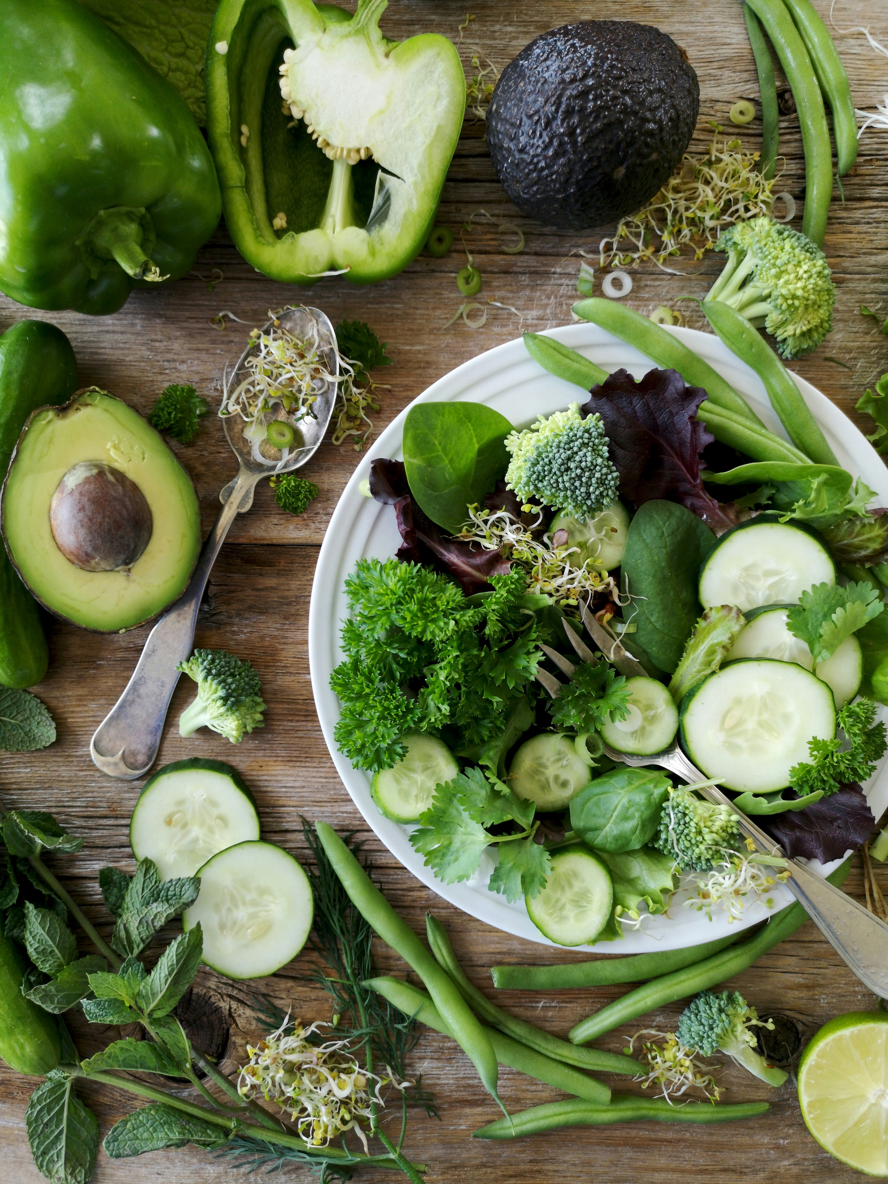 wooden table covered with green vegetables and a plate with broccolli, avocado, bell peppers, snow peas with a metal fork