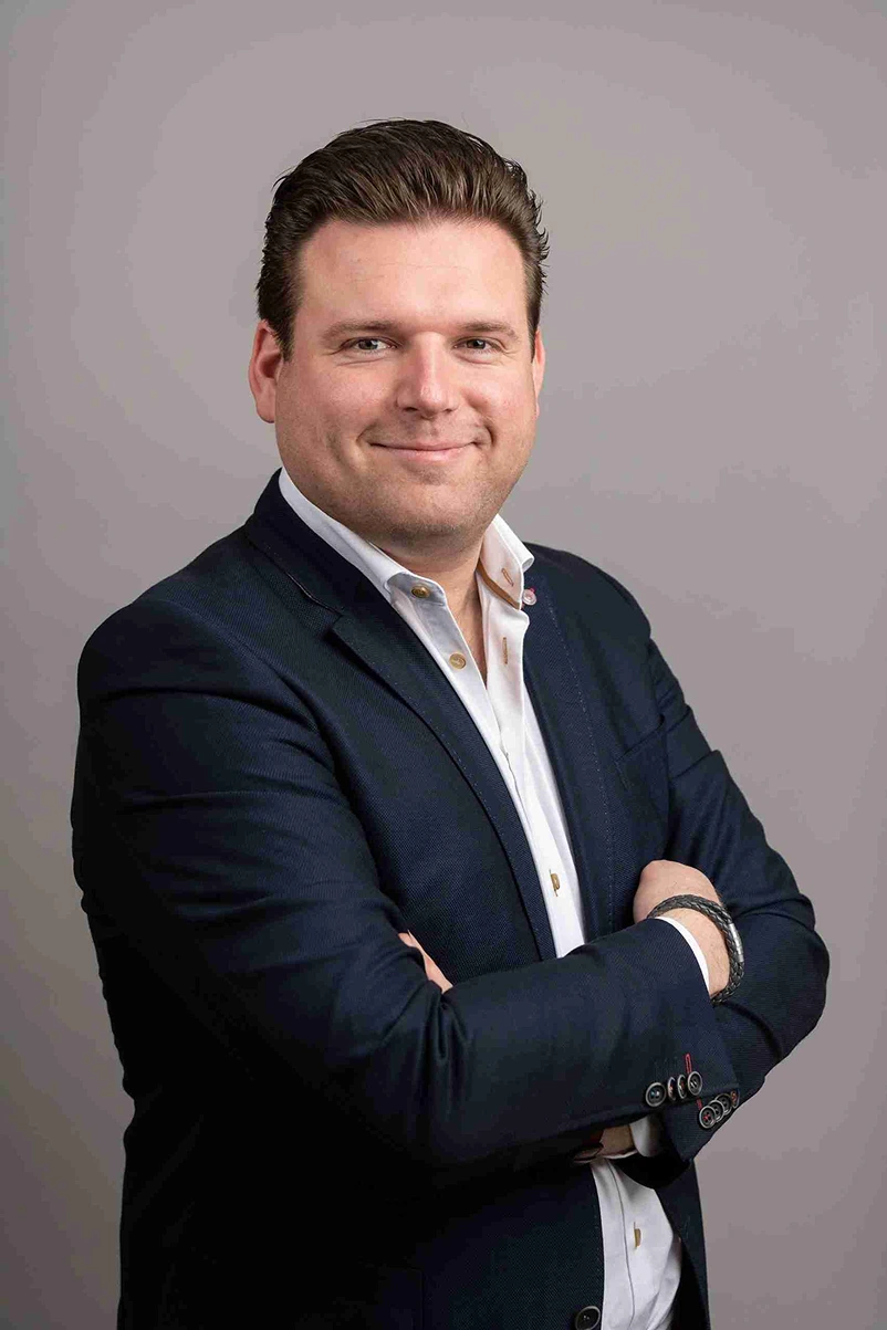 Smiling man with slicked-back hair wearing a navy blazer and white shirt, standing with arms crossed against a plain gray background.