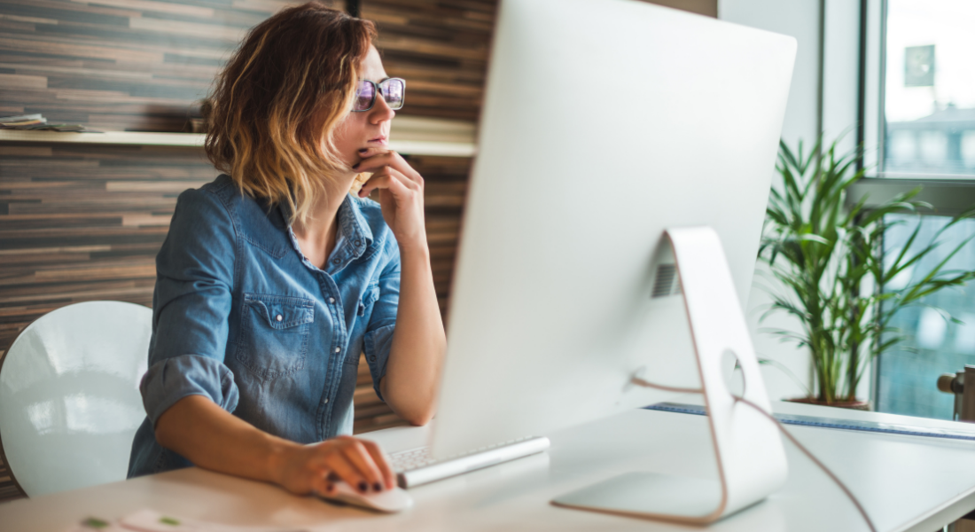 Thoughtful woman in glasses sitting at desk, looking stressed while working on computer in modern office
