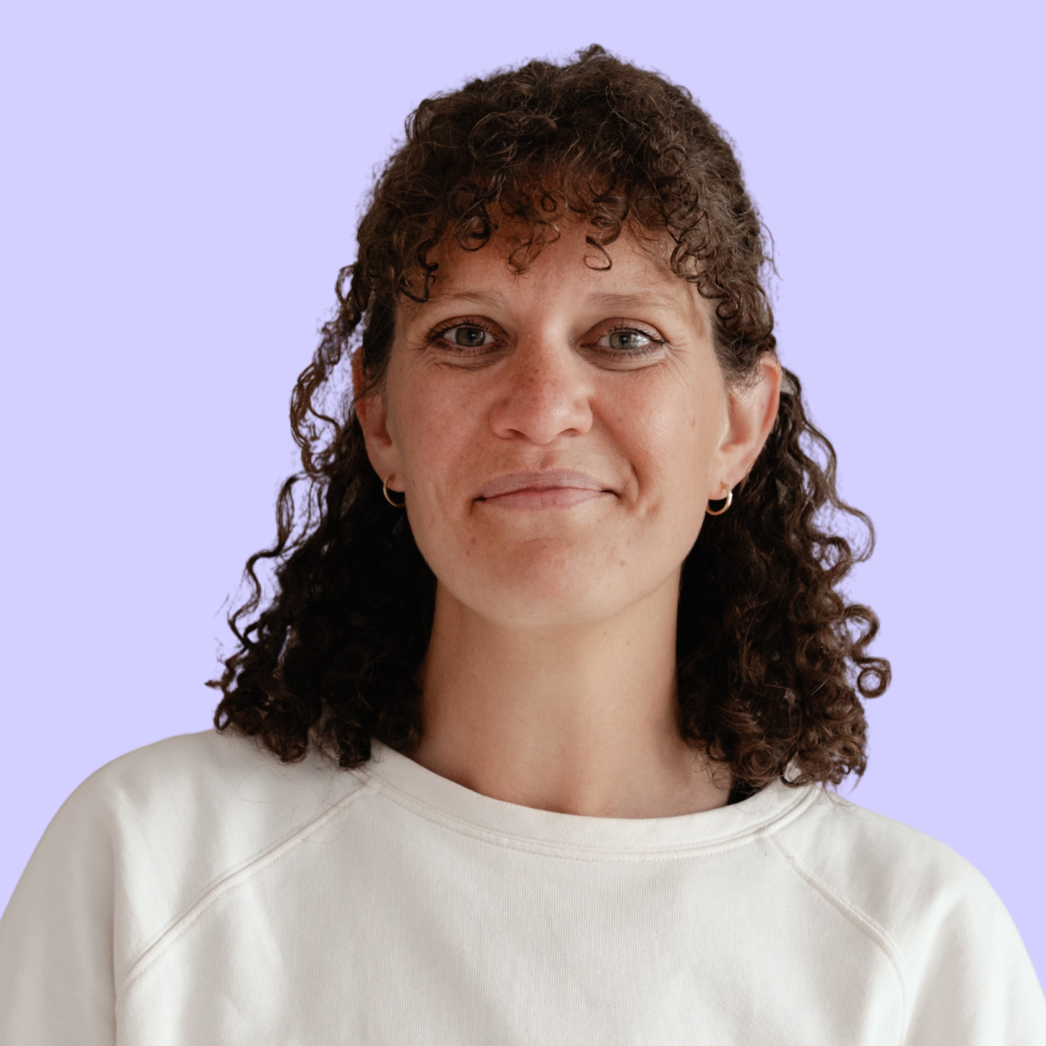 Sabine Antweiler schaut in die Kamera und lächelt Zähne zeigend. Diese Person hat langes Haar, welches sie zu einen Dutt zusammengebunden hat und trägt ein schwarzes Oberteil. Der Hintergrund ist rosa.