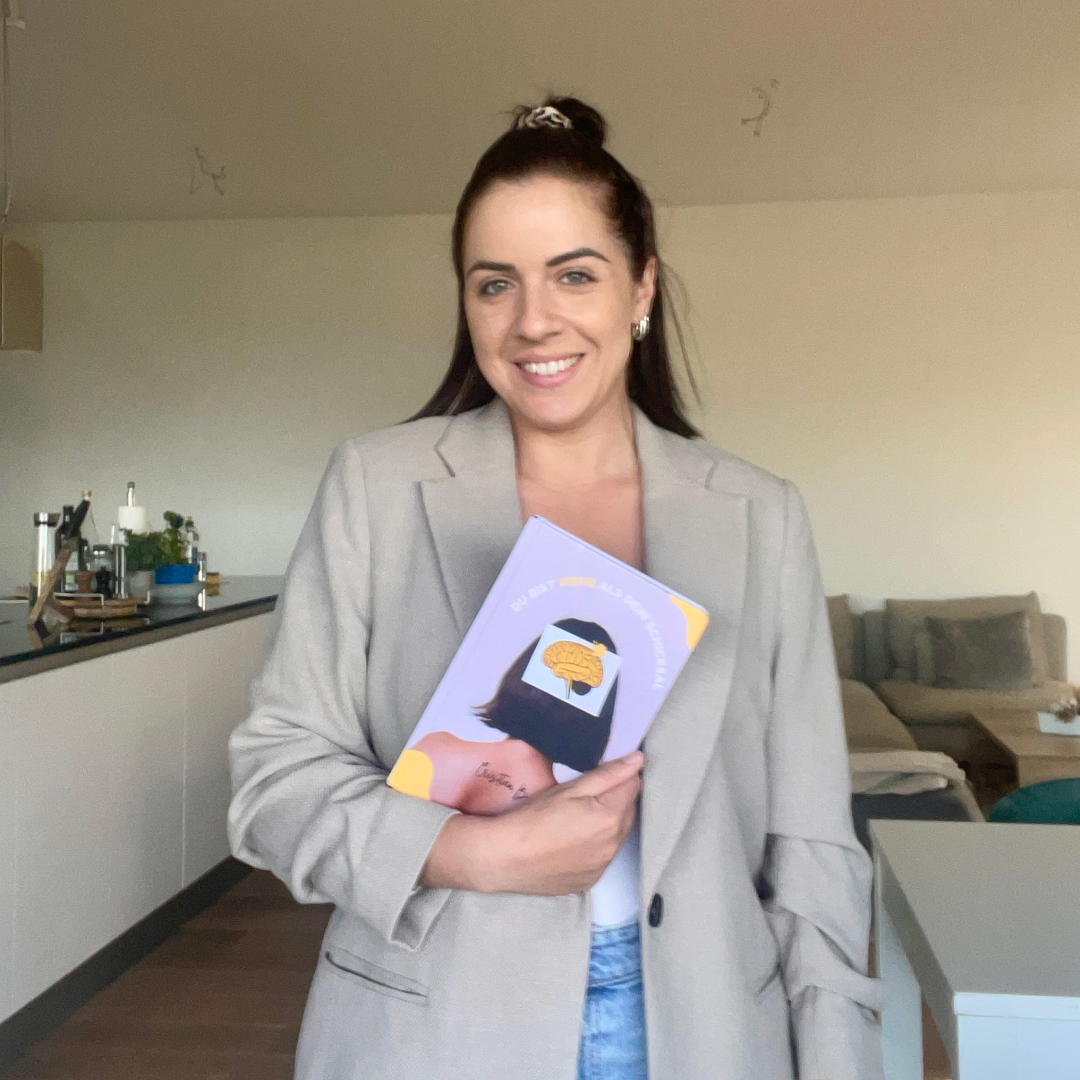 Eine Frau im beigen Blazer hält ein Buch und lächelt.