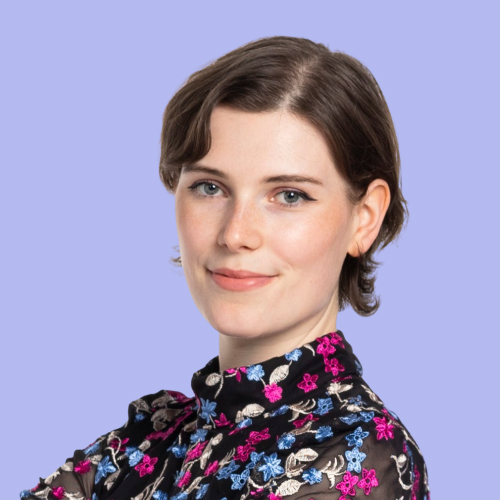 Katharina hat kurze, lockige, braune Haare und trägt ein Oberteil mit Blumen. Der Hintergrund ist lila. 