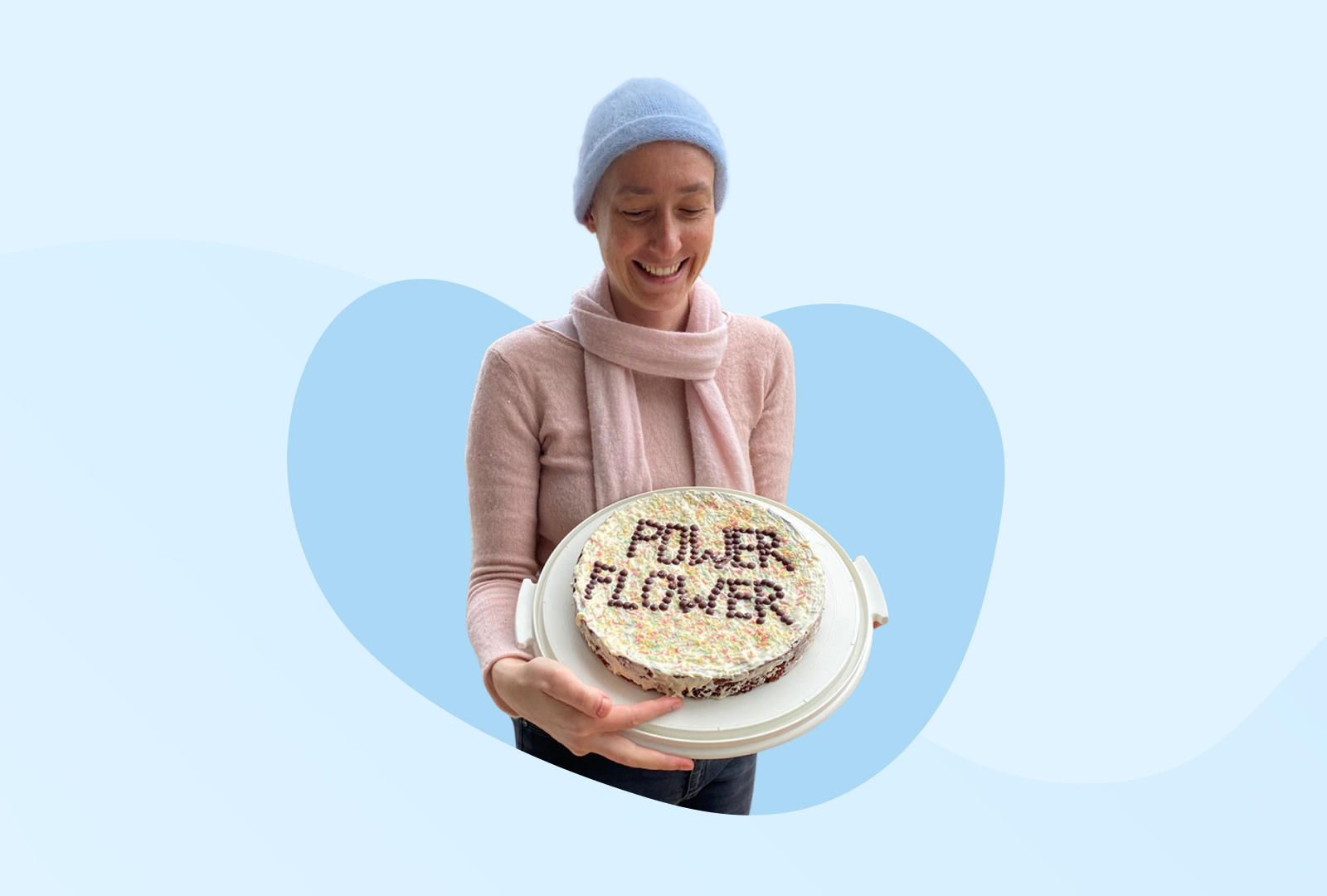 Steffi trägt eine blaue Mütze und hält lächelnd eine Torte mit der Aufschrift Power-Flower in den Händen.