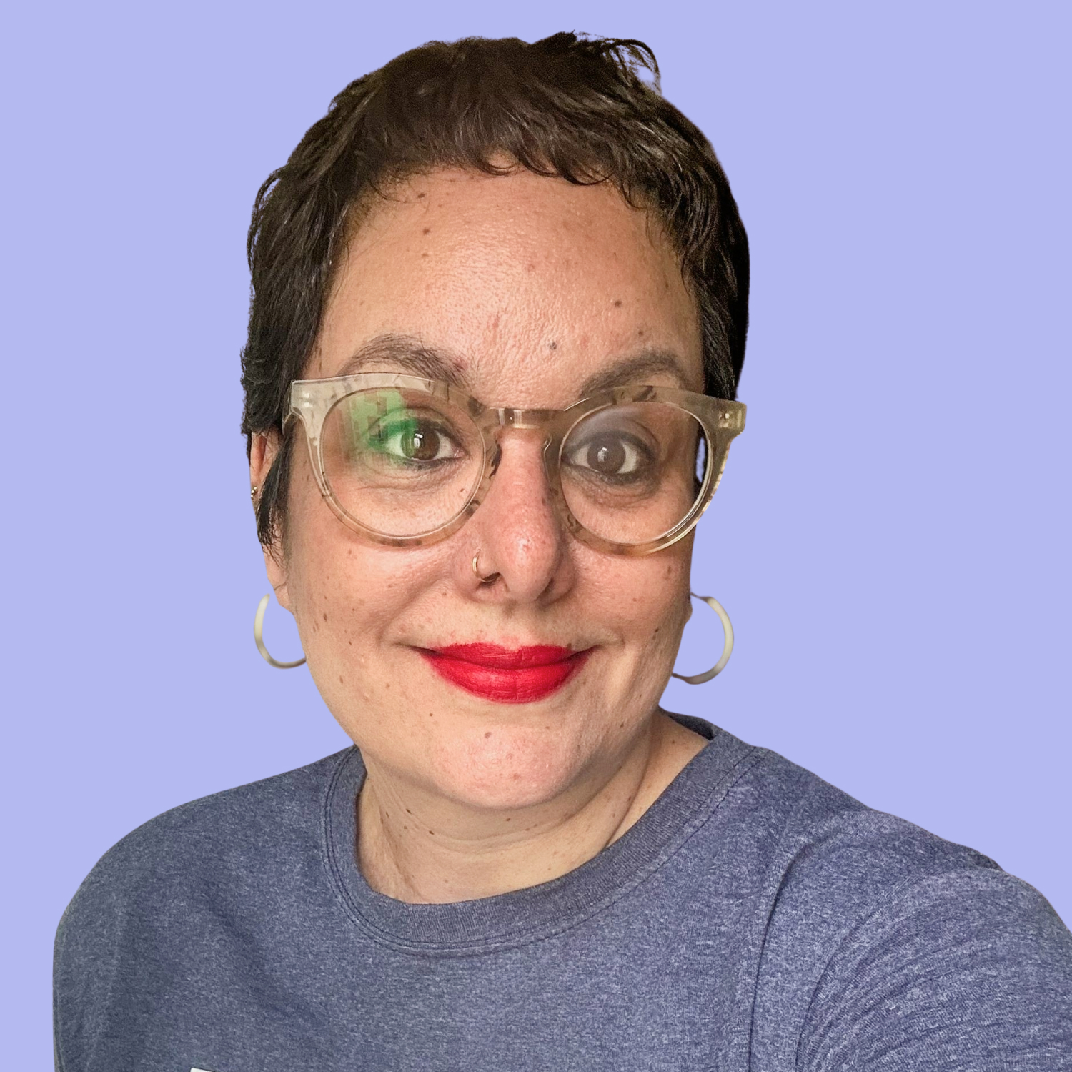 Porträt von Jasmin mit kurzem dunklem Haar, heller Brille, roten Lippen und silbernen Creolen vor einem hellvioletten Hintergrund.