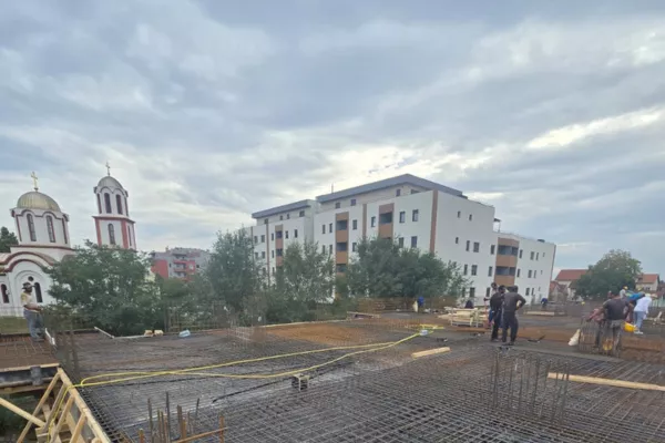 Construction site with steel rebar framework and workers, with residential buildings and a church in the background under a cloudy sky.