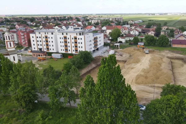 Aerial view of a suburban area with a large white building under construction next to a cleared dirt plot and surrounded by green trees and houses.