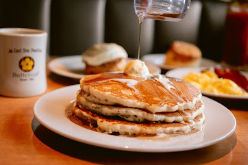 Stack of pancakes with butter and syrup being poured on top, served with scrambled eggs and biscuits in the background.