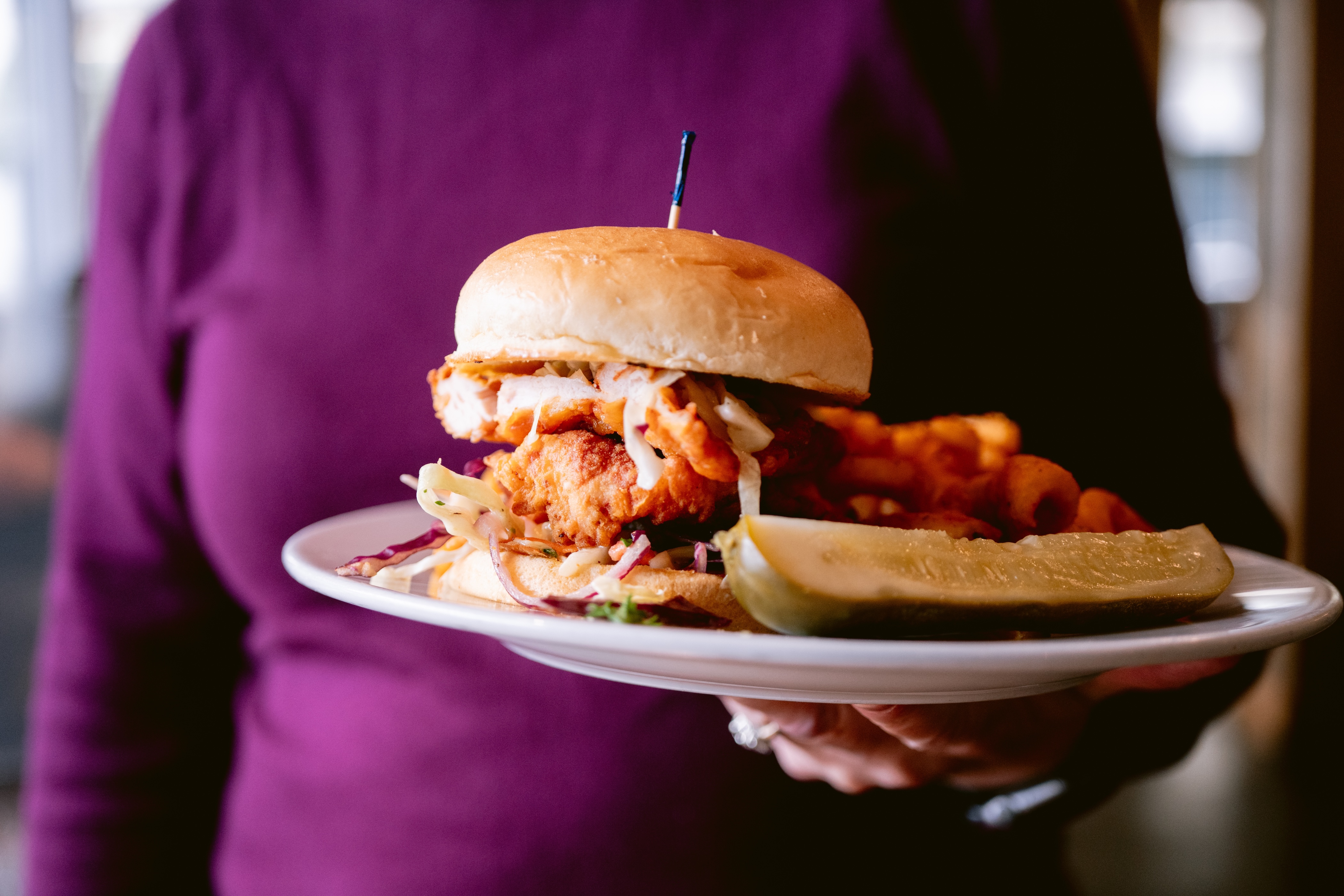 Person in purple shirt holding a plate with a fried chicken sandwich, pickle spear, and curly fries.