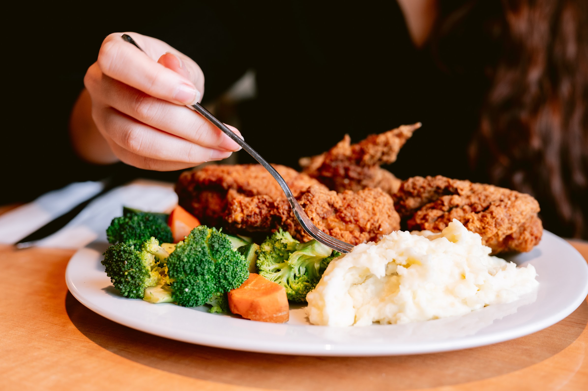 Hand holding a fork picking up broccoli from a plate with fried chicken and mashed potatoes.