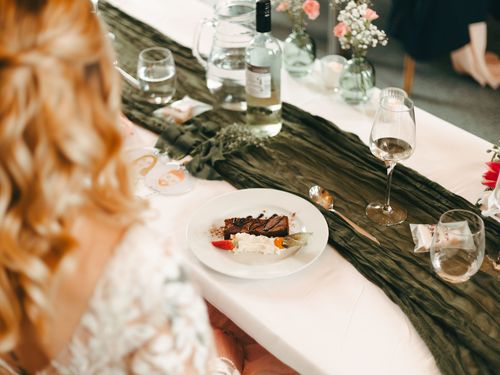 A person garnishes a plate with herbs. The dish includes a piece of meat, a roasted carrot, mashed potatoes in a small bowl, and possibly other items. The plate rests on a tray, suggesting a restaurant or professional kitchen setting.