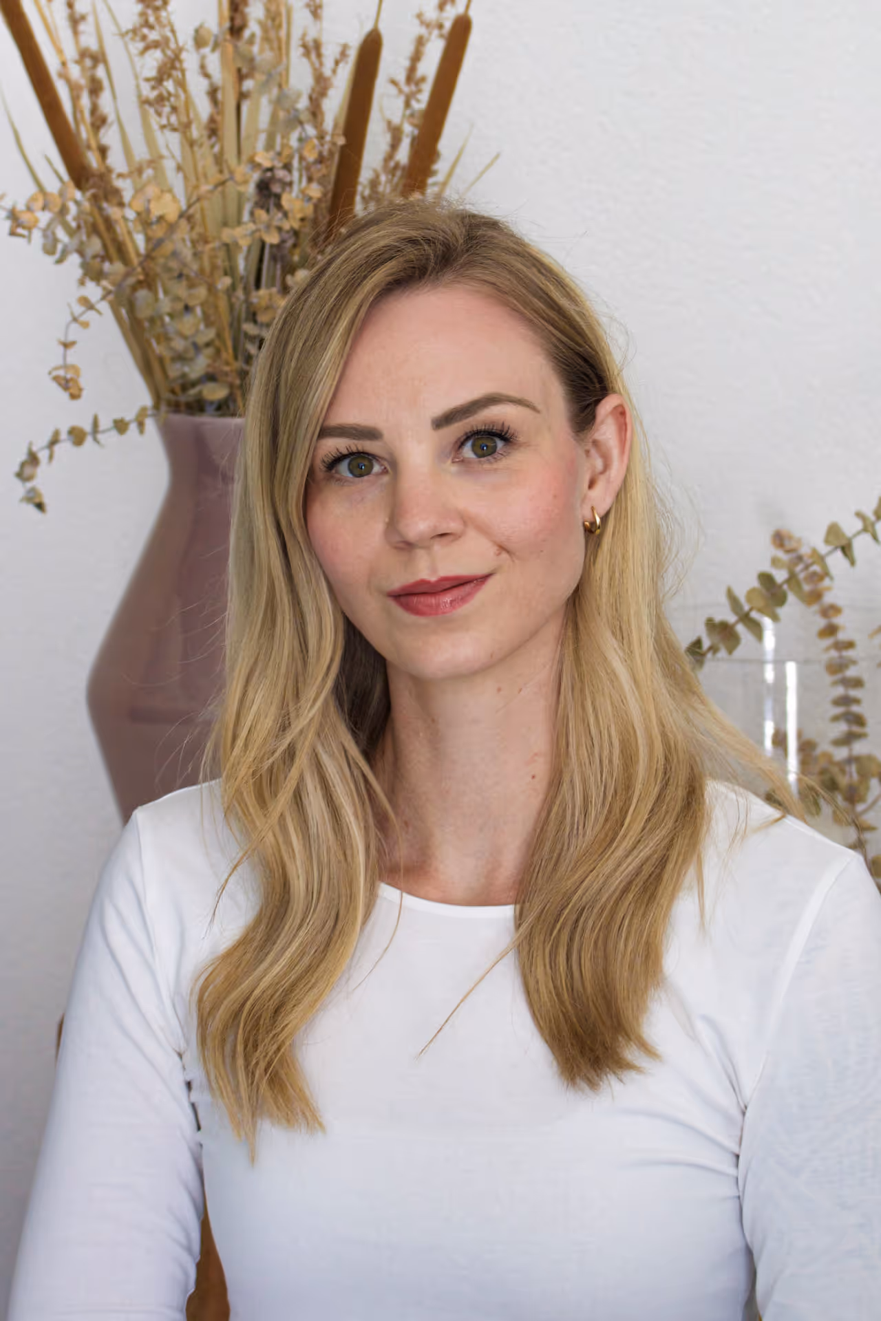 Porträt einer lächelnden Frau (Anja Steiner) mit langen blonden Haaren und weißem Oberteil vor einer Vase mit getrockneten Pflanzen.