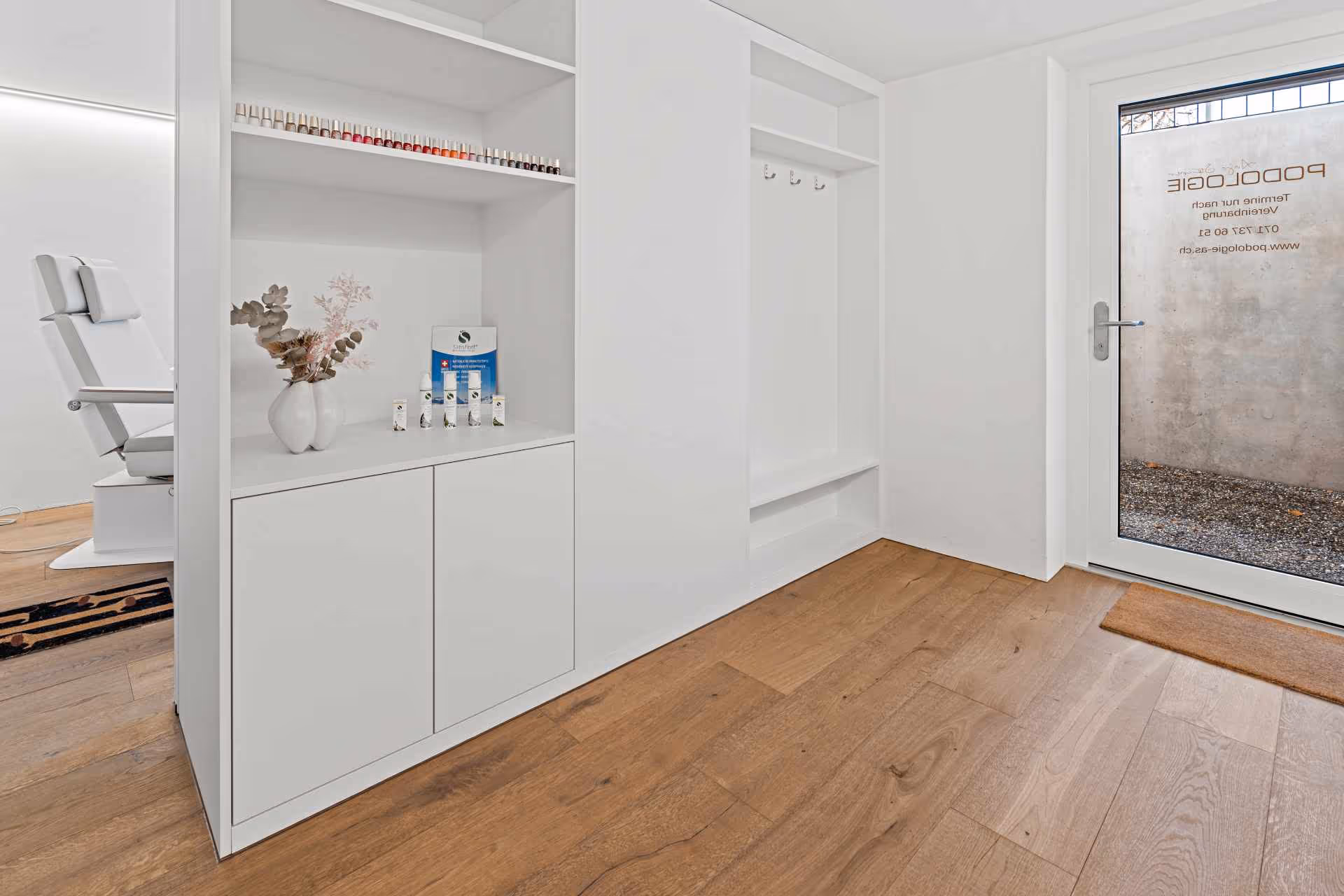 Heller Eingangsbereich einer Podologie-Praxis mit weißen Regalen, Nagellackfläschchen, Vase mit Trockenblumen und Liegestuhl im Behandlungsraum.