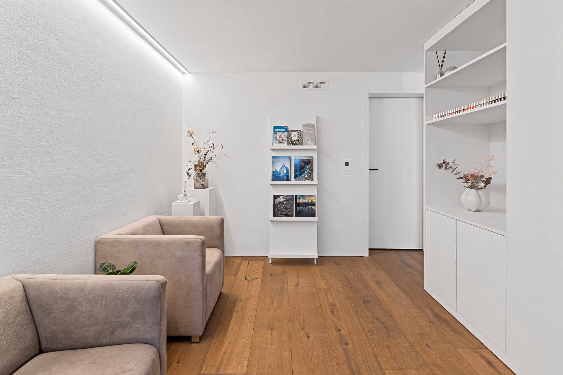 Hell eingerichteter Wartebereich mit beigen Sesseln, Holzboden, weißen Wänden, einem Regal mit Magazinen und Blumenvasen.