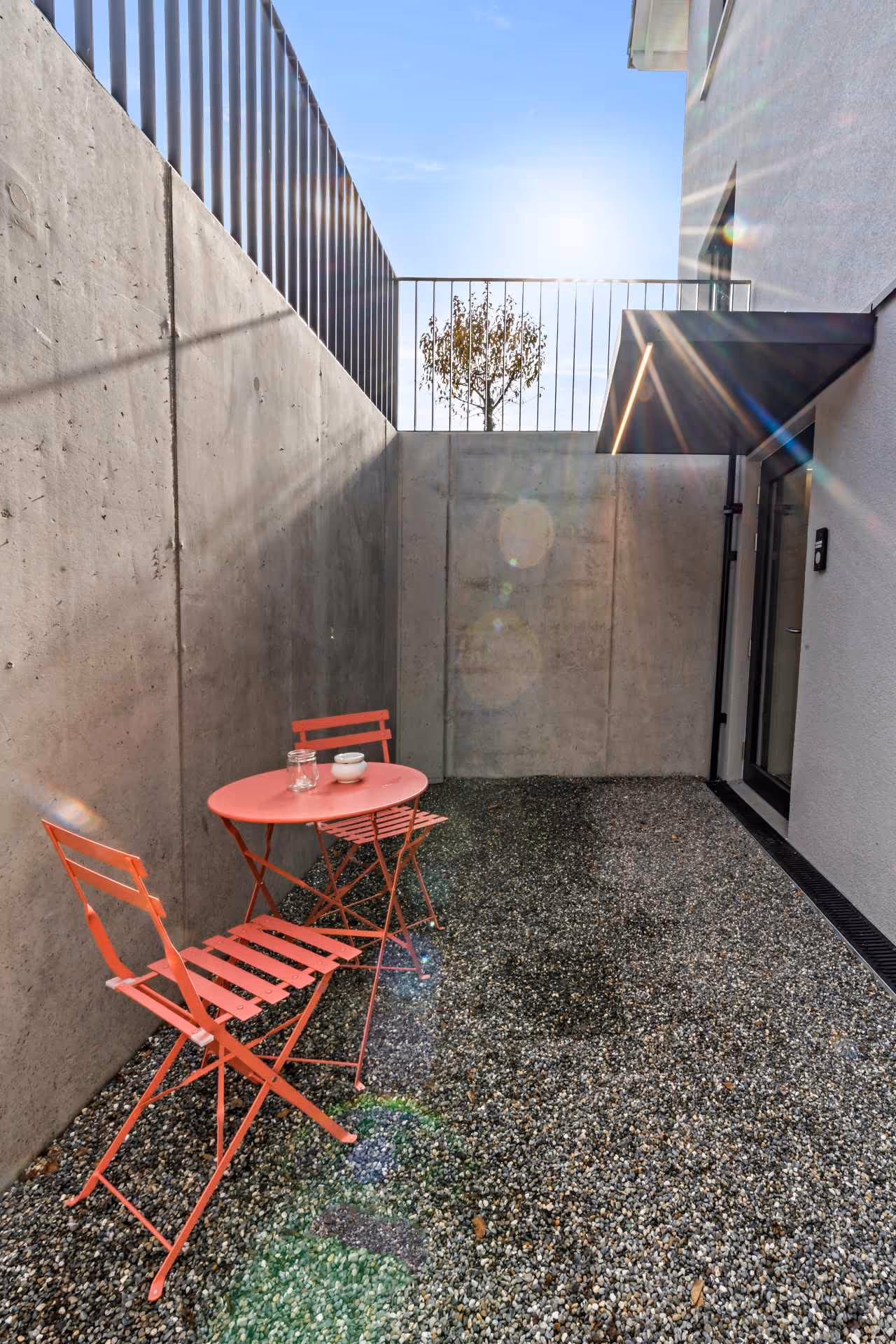 Kleiner Sitzbereich mit rotem Metalltisch und zwei Stühlen auf Kiesboden neben einer Betonwand, Sonnenlicht und Baum hinter einem Geländer sichtbar.
