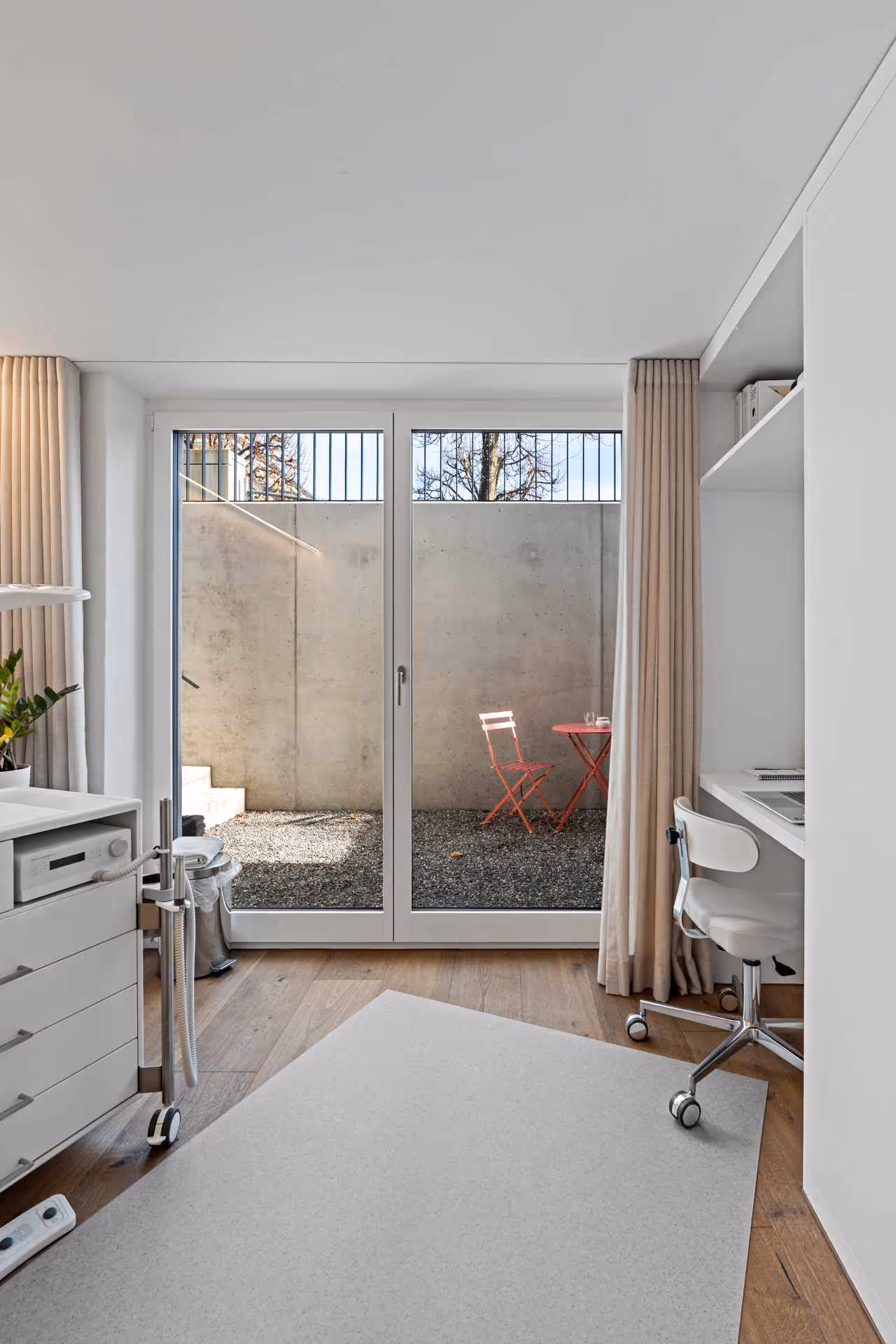 Heller, moderner Raum mit Holzparkett, weissem Bürostuhl, Schreibtisch, Schränken und grossen Glastüren, die auf einen kleinen Innenhof mit rotem Klapptisch und Stuhl führen.