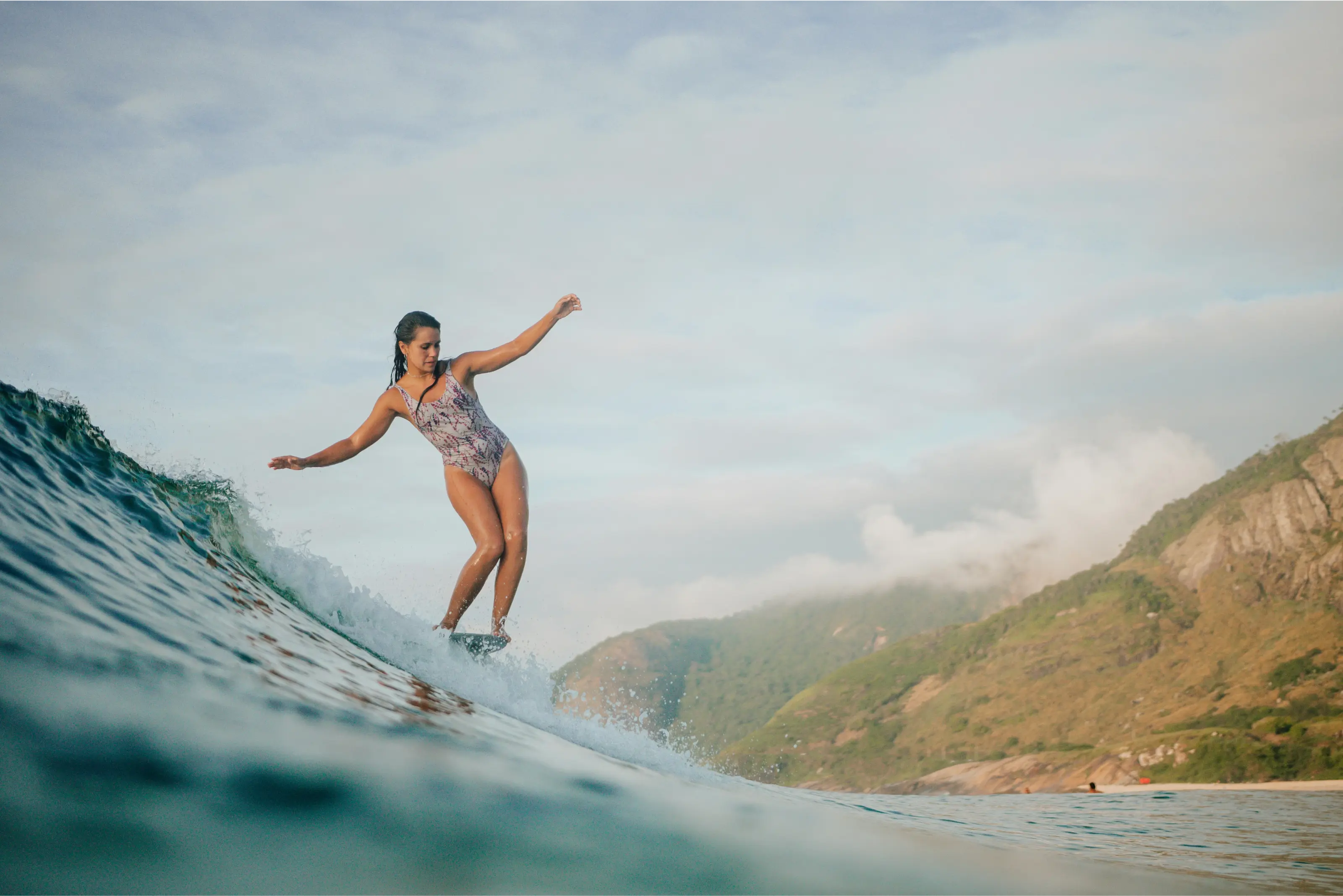 Woman surfing on a wave near a rocky, green hillside under a cloudy sky.