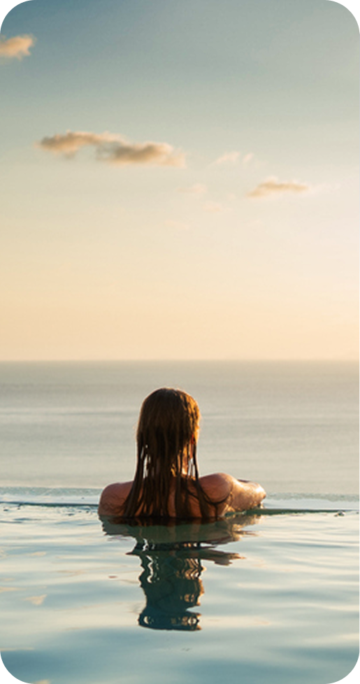 Person with wet hair relaxing in an infinity pool overlooking a calm ocean at sunset.