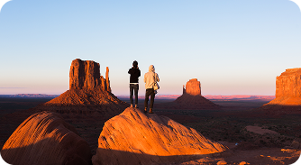 Two people standing on a rock overlooking Monument Valley during sunset.