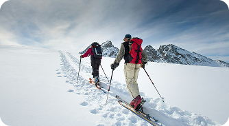 Two skiers ascending a snowy mountain with clear sky and rocky peaks in the background.