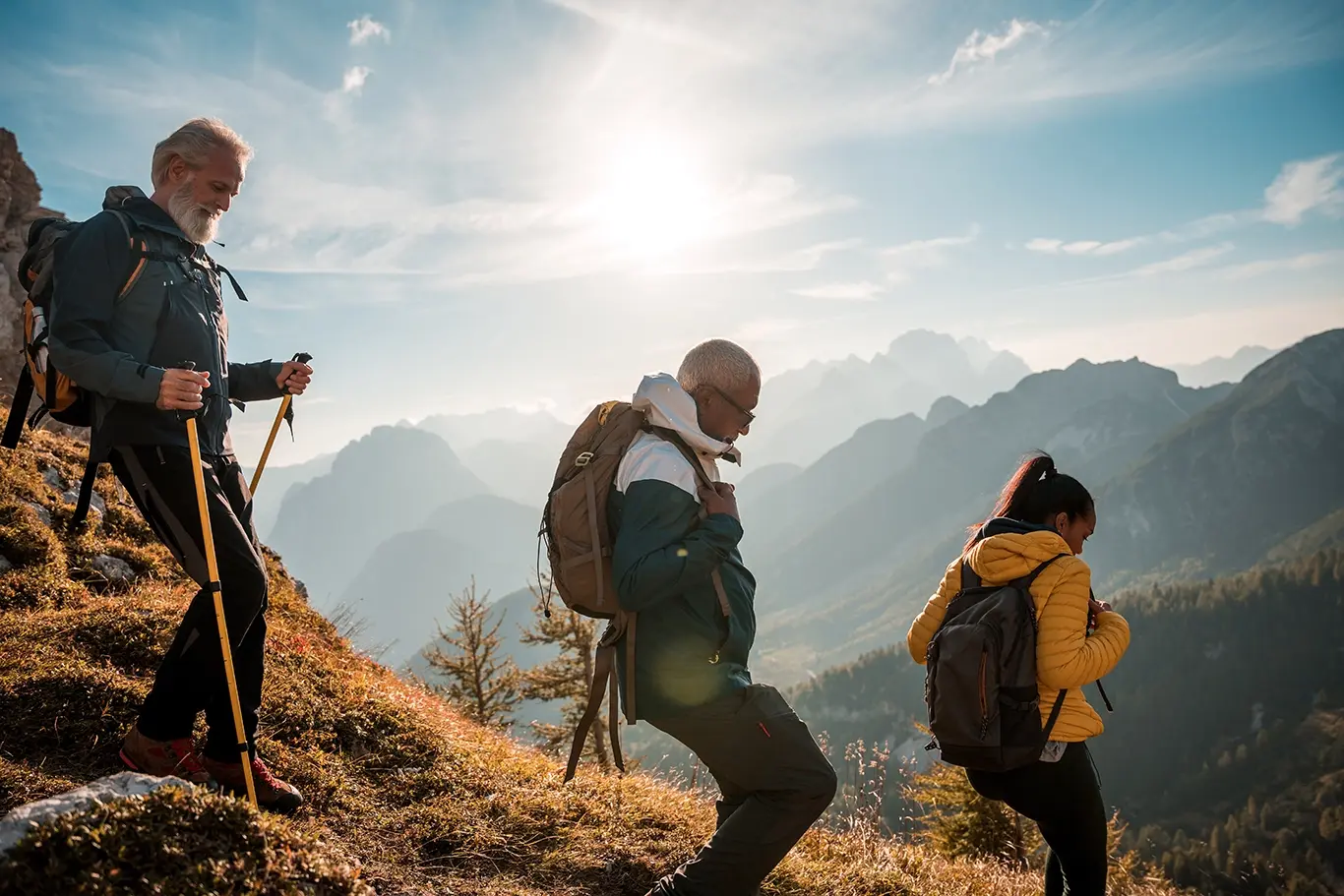 Three hikers with backpacks descend a mountain trail in sunny, rugged terrain with distant peaks.