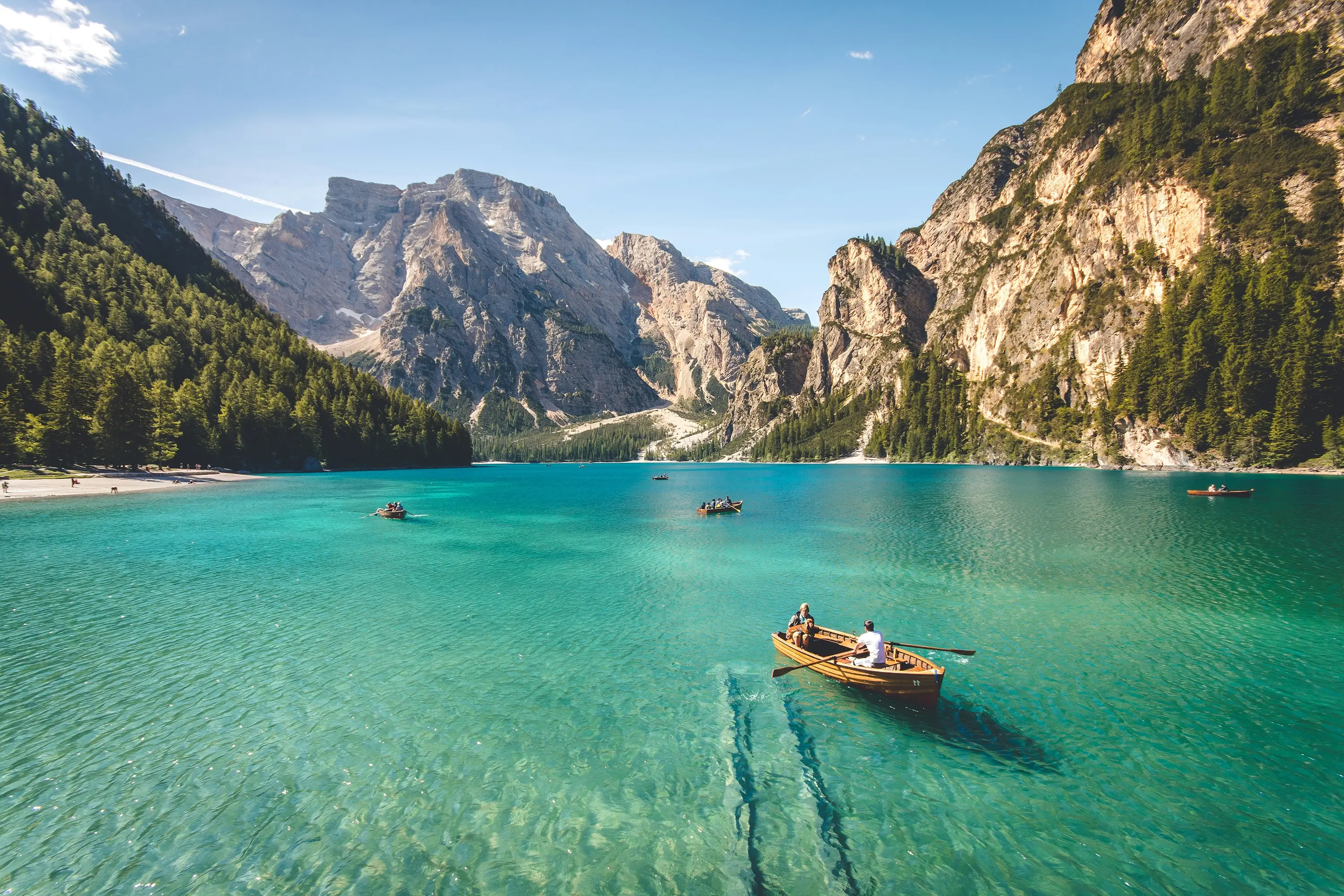People rowing wooden boats on clear turquoise lake surrounded by forested mountains under blue sky.