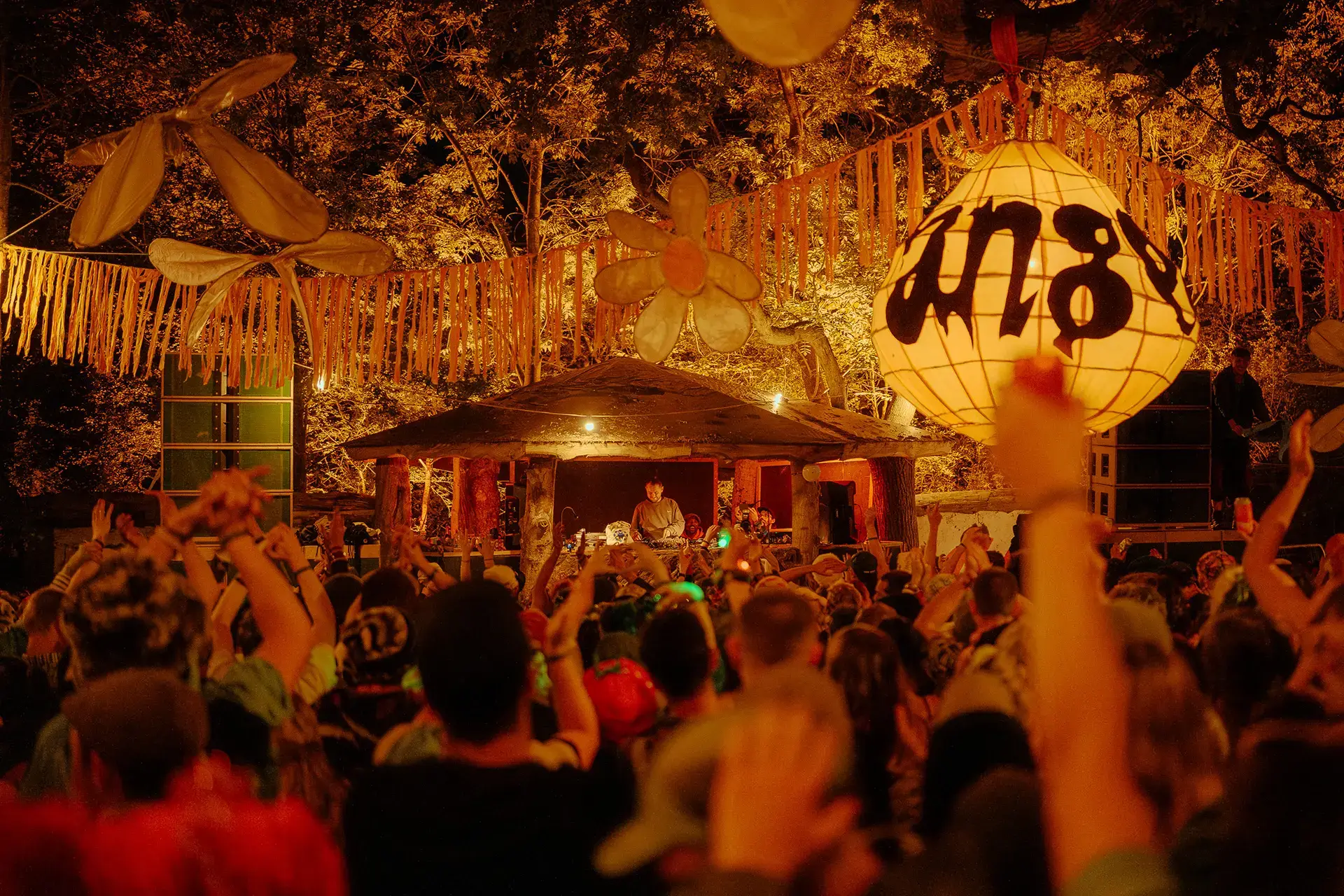 Crowd dancing beneath warm lantern lighting at An Experience Festival, with a DJ performing on a woodland stage at night.