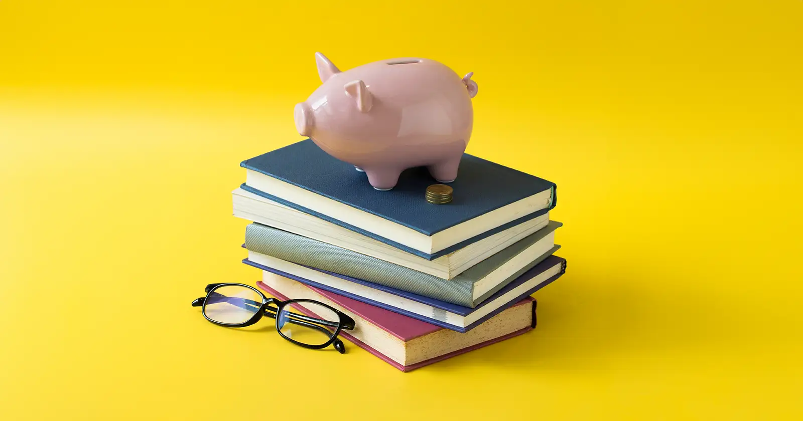 Pink ceramic piggy bank on top of a stack of four closed books with a pair of black eyeglasses beside them against a yellow background.