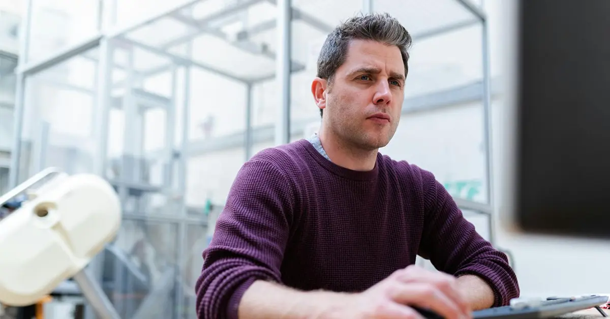 A person in a purple sweater holds a computer mouse while dubiously looking at a computer screen