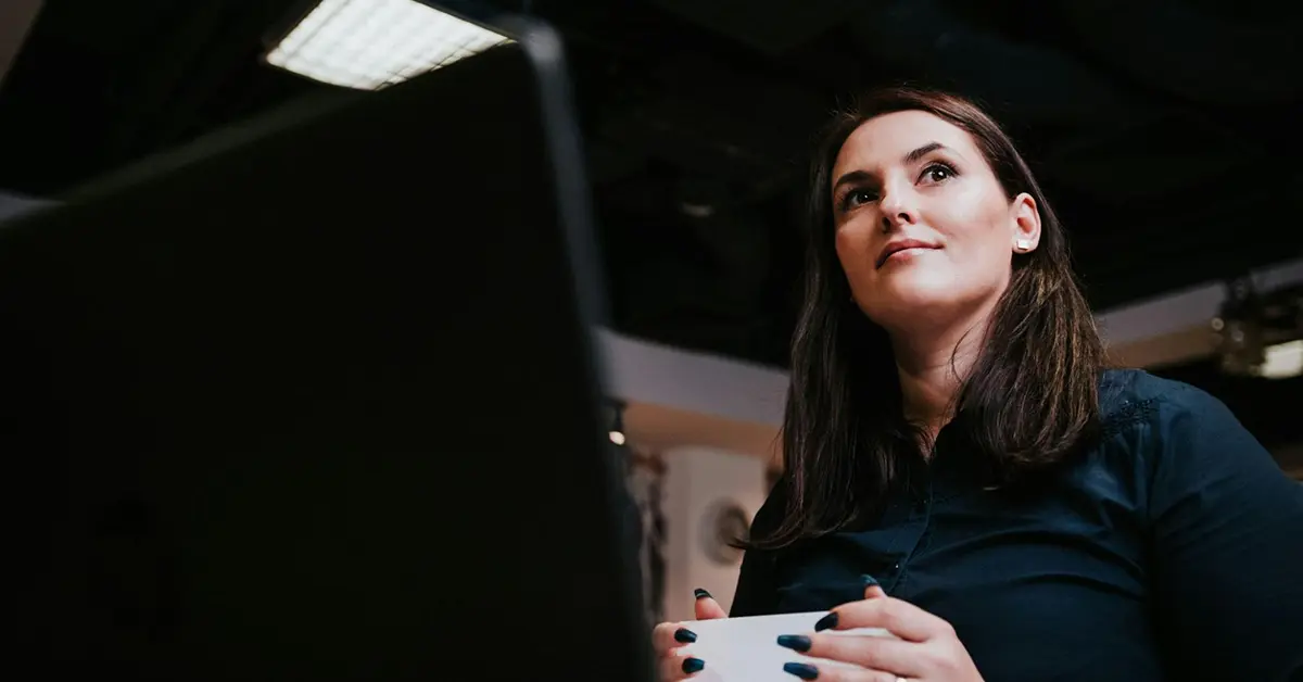 Low-angle shot of a person looking up from their laptop while holding a stack of papers.