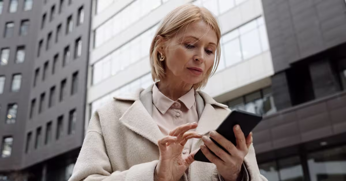 A woman in white coat looks at her smartphone while walking in front of a tall building