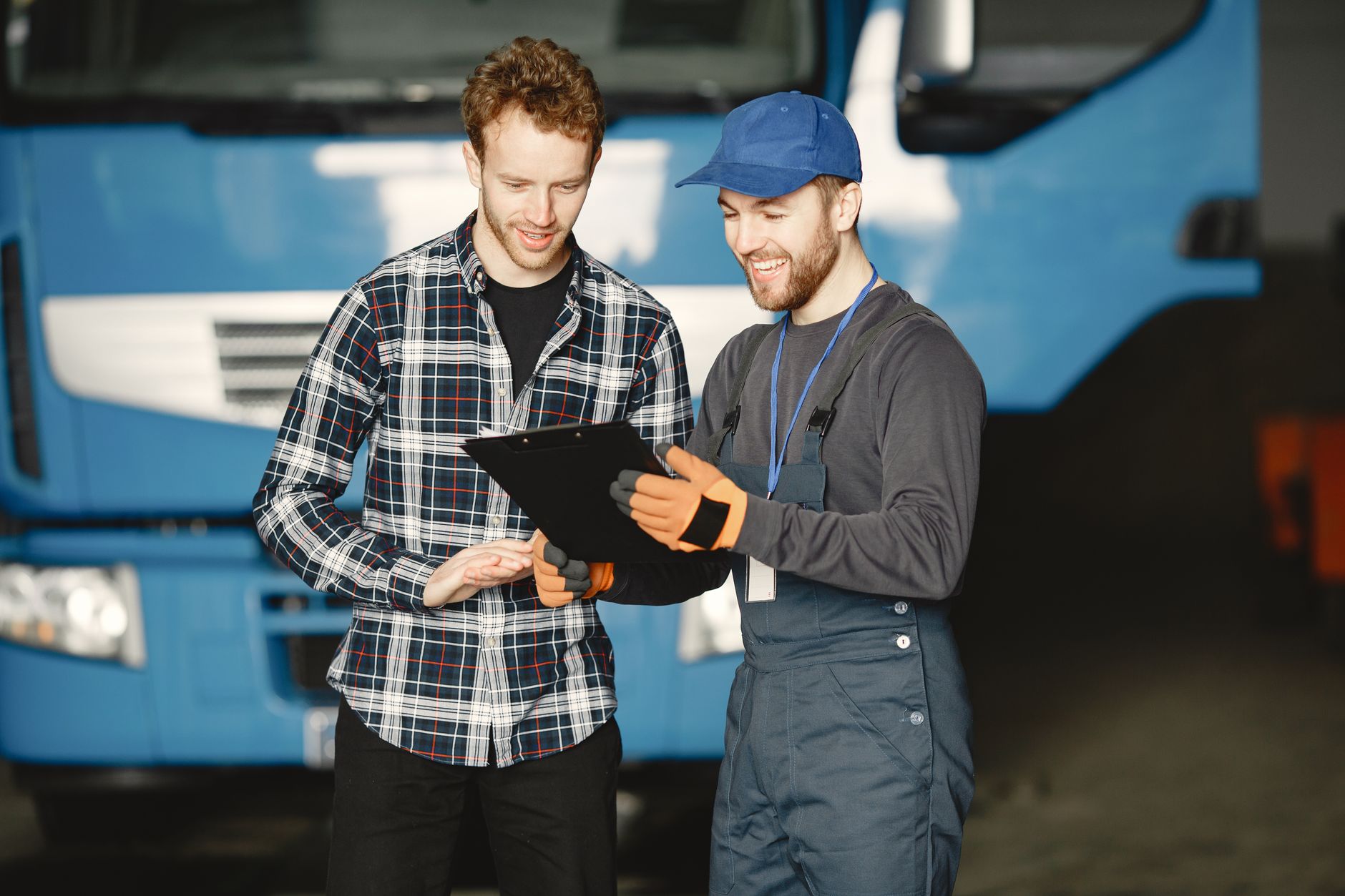 Two men discussing truck repairs in a garage: one in mechanic uniform, the other in a plaid shirt.