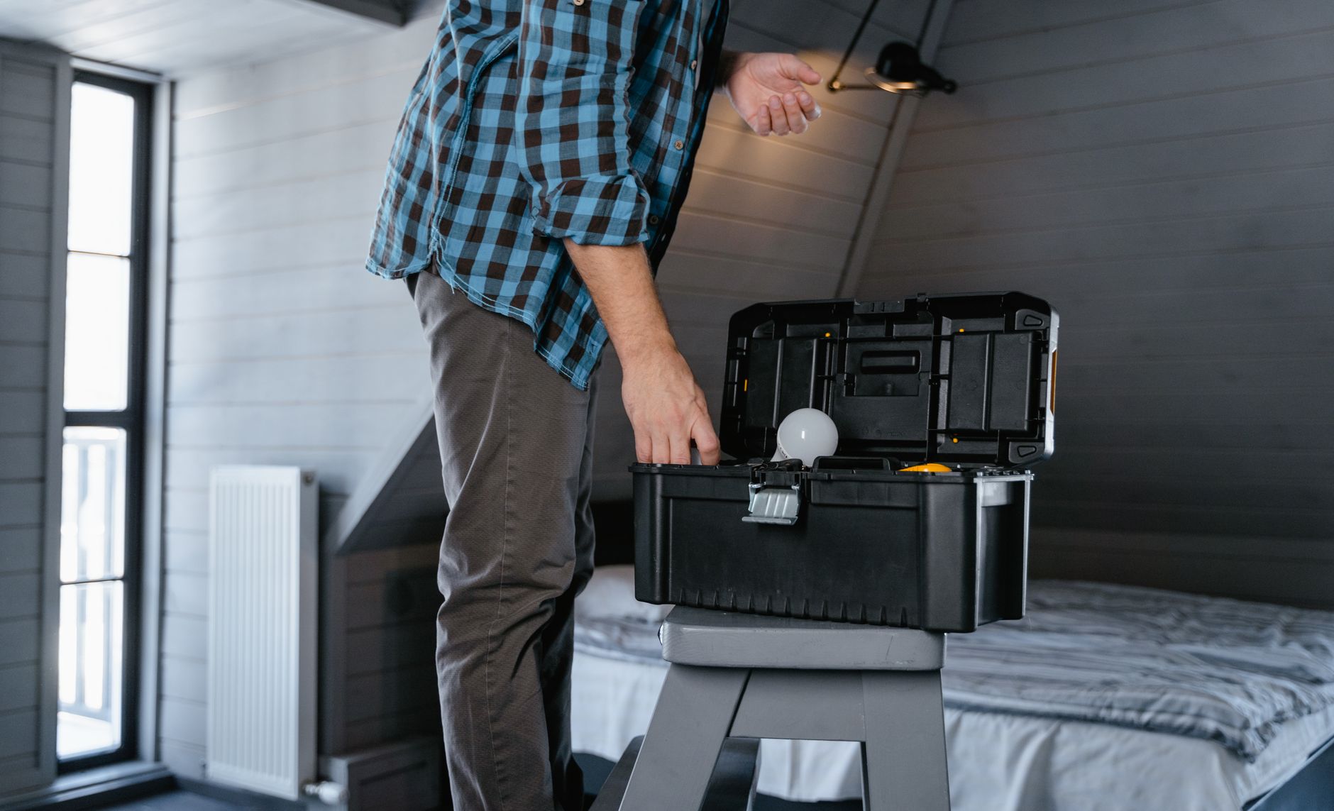 Person in plaid shirt fixing a light fixture using a toolbox in a rustic attic interior.