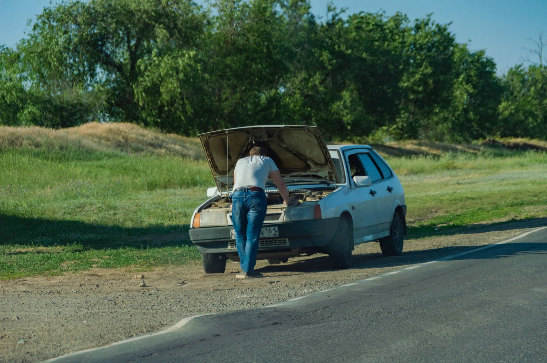 Man standing at a parked blue car with the hood open on a country road