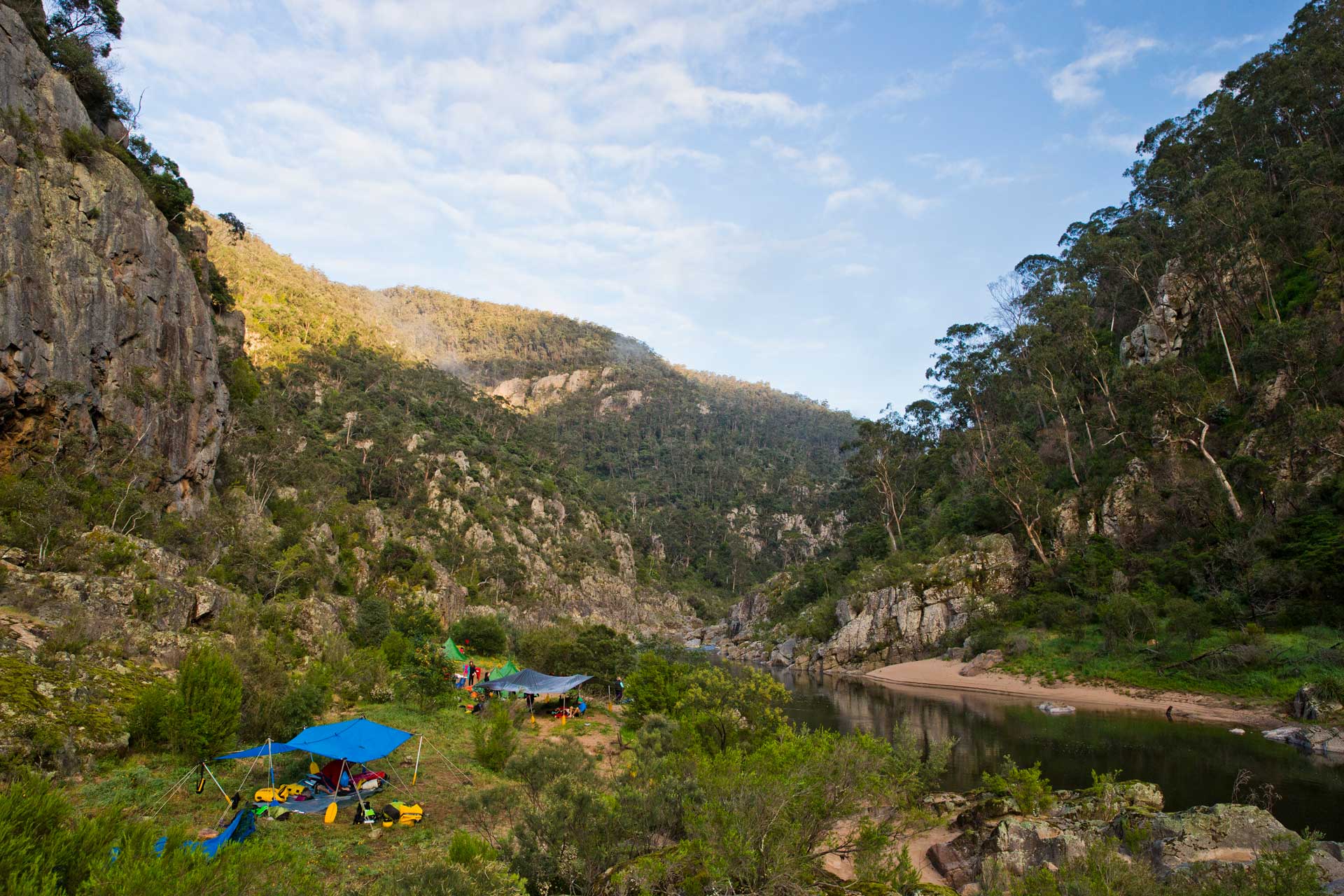 Camping site with blue tarps and kayaks beside a calm river surrounded by rocky hills and forested landscape.