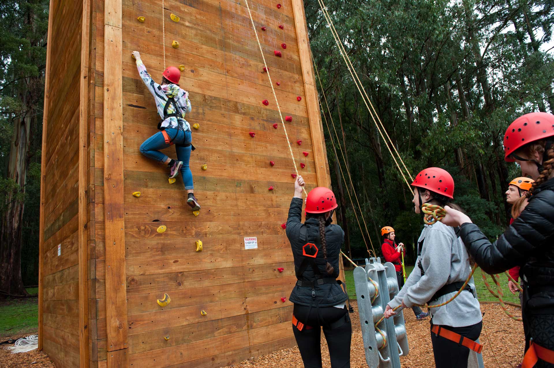 Person in red helmet climbs wooden wall with colourful holds while group in safety gear waits below with belay ropes.