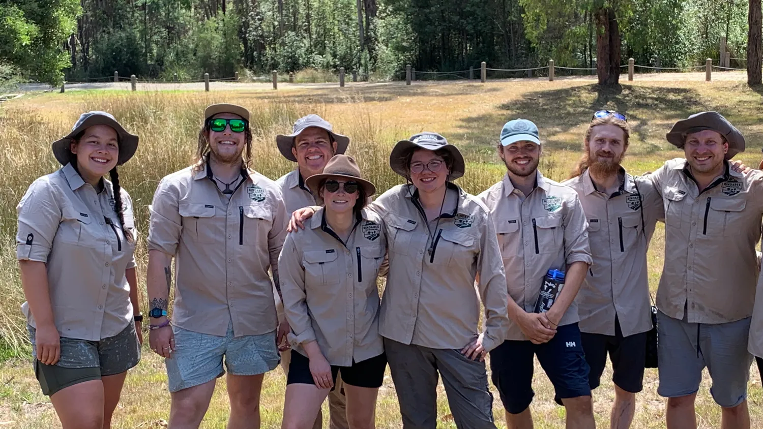 Eight people in matching khaki uniforms and hats posing together outdoors in a grassy field with trees in the background.