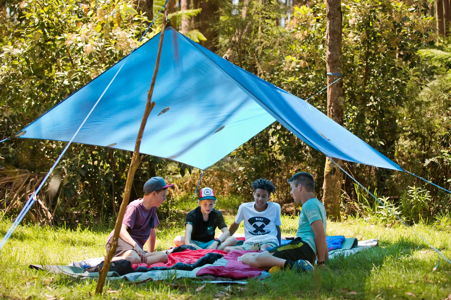Four young people sit under a blue tarp shelter on blankets in a wooded area, having a casual outdoor gathering.