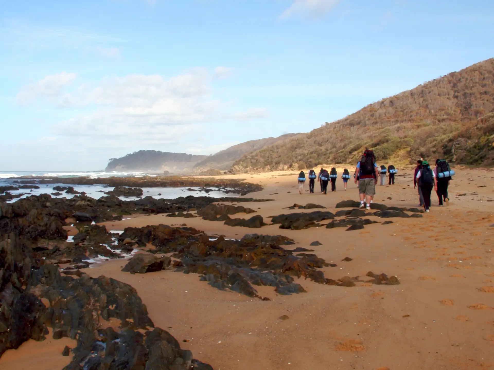 Group walking on sandy beach with rocky outcrops, hills in background under partly cloudy sky at low tide.