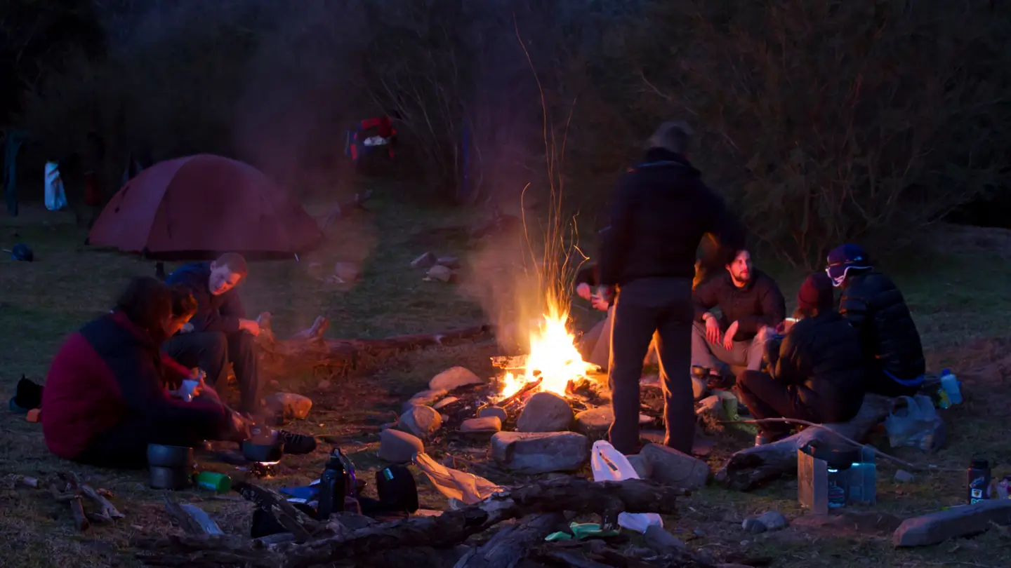 Campers gathered around a glowing campfire at dusk with tents visible in the background amongst bushland vegetation.