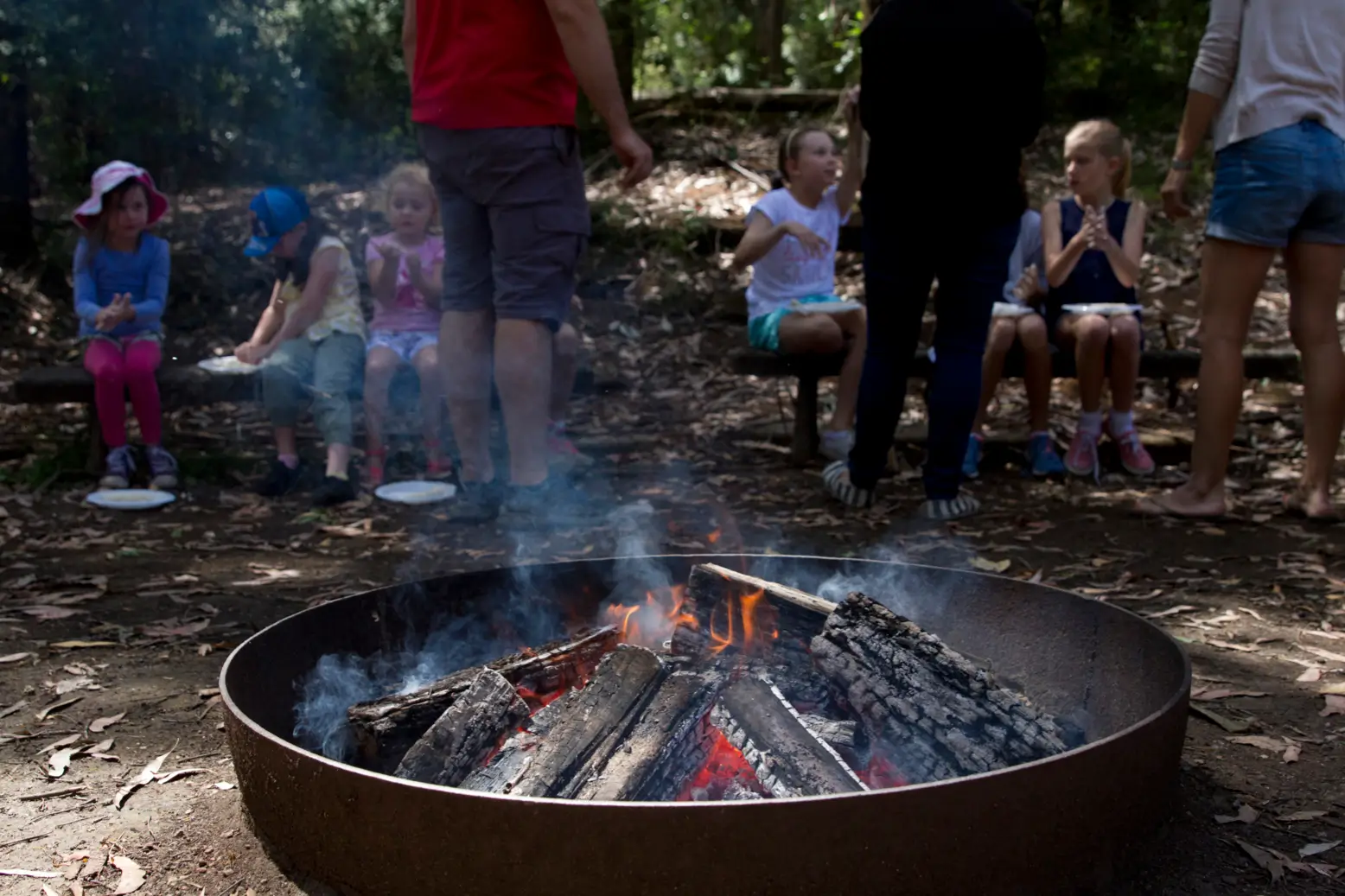 Children seated on benches watching a campfire burn in a metal fire ring during an outdoor forest activity with supervising adults.