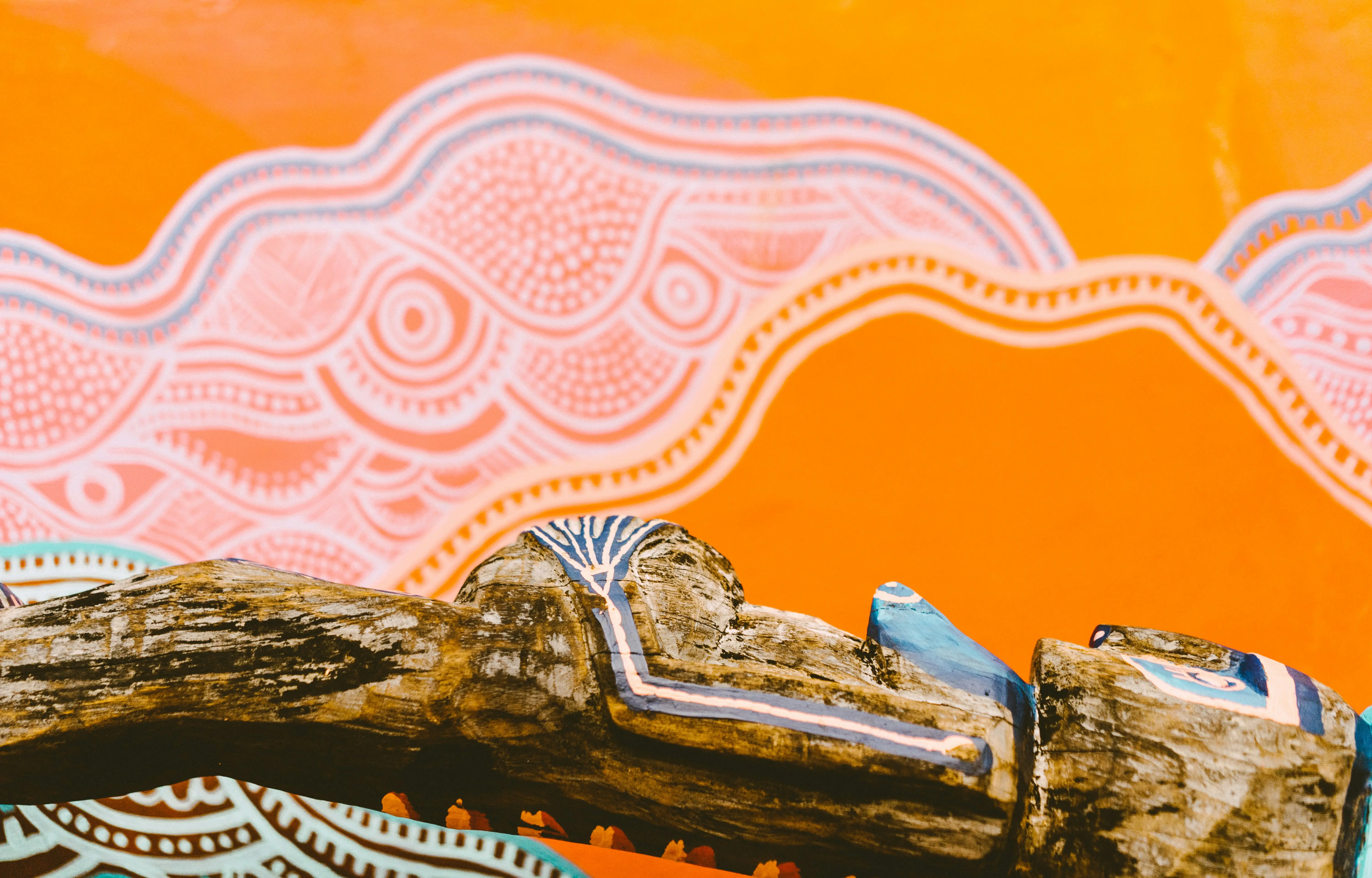 A weathered wooden branch painted with Indigenous Australian art patterns displayed against an orange backdrop featuring traditional designs.