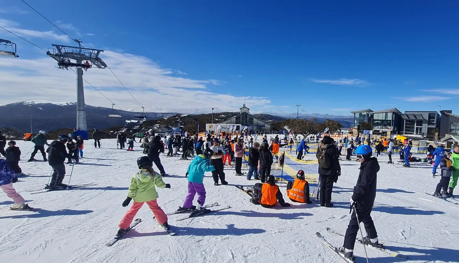 A crowded ski resort scene with skiers and snowboarders on snowy slopes, chairlifts, and mountain buildings under blue skies.