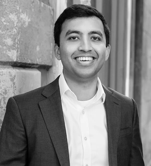 Smiling man in a suit jacket and white shirt standing against a stone wall.