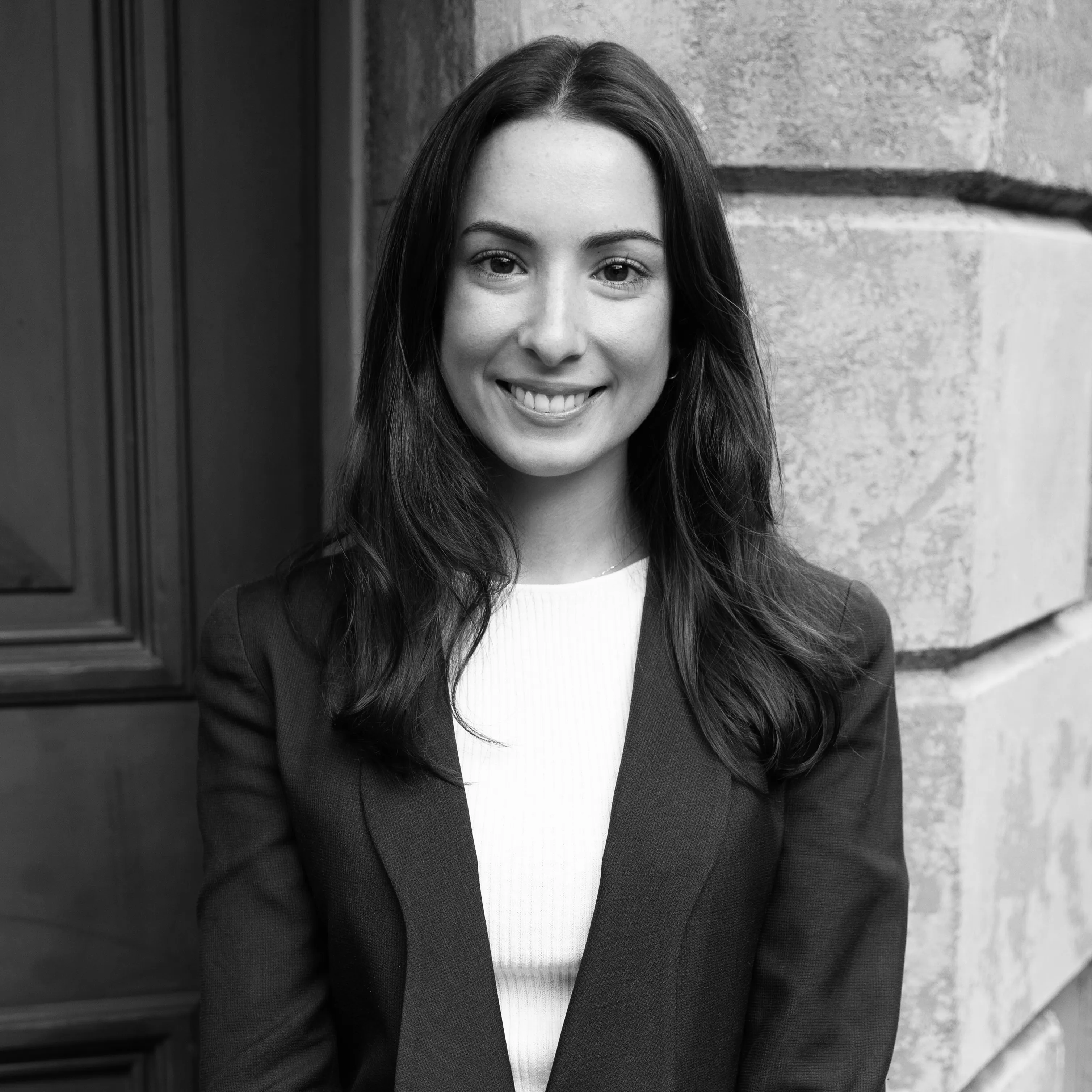 Smiling woman with long dark hair wearing a blazer and white top standing in front of a stone wall and wooden door.
