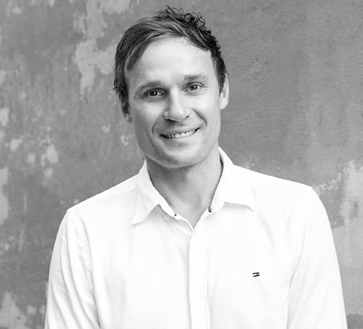 Smiling man with short hair wearing a white button-up shirt against a textured gray wall.