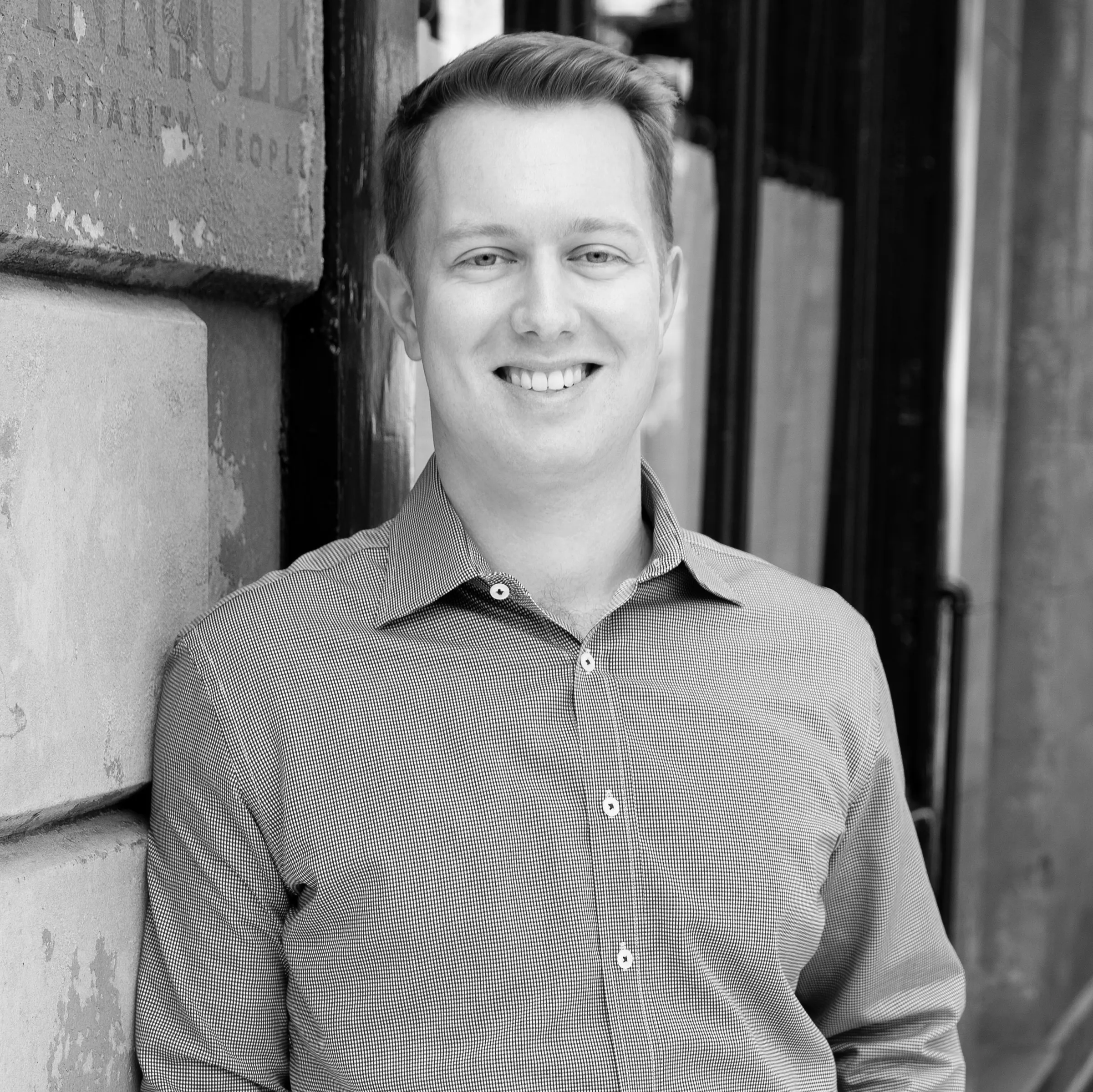 Smiling man in a button-up shirt leaning against an outdoor stone wall in an urban setting.