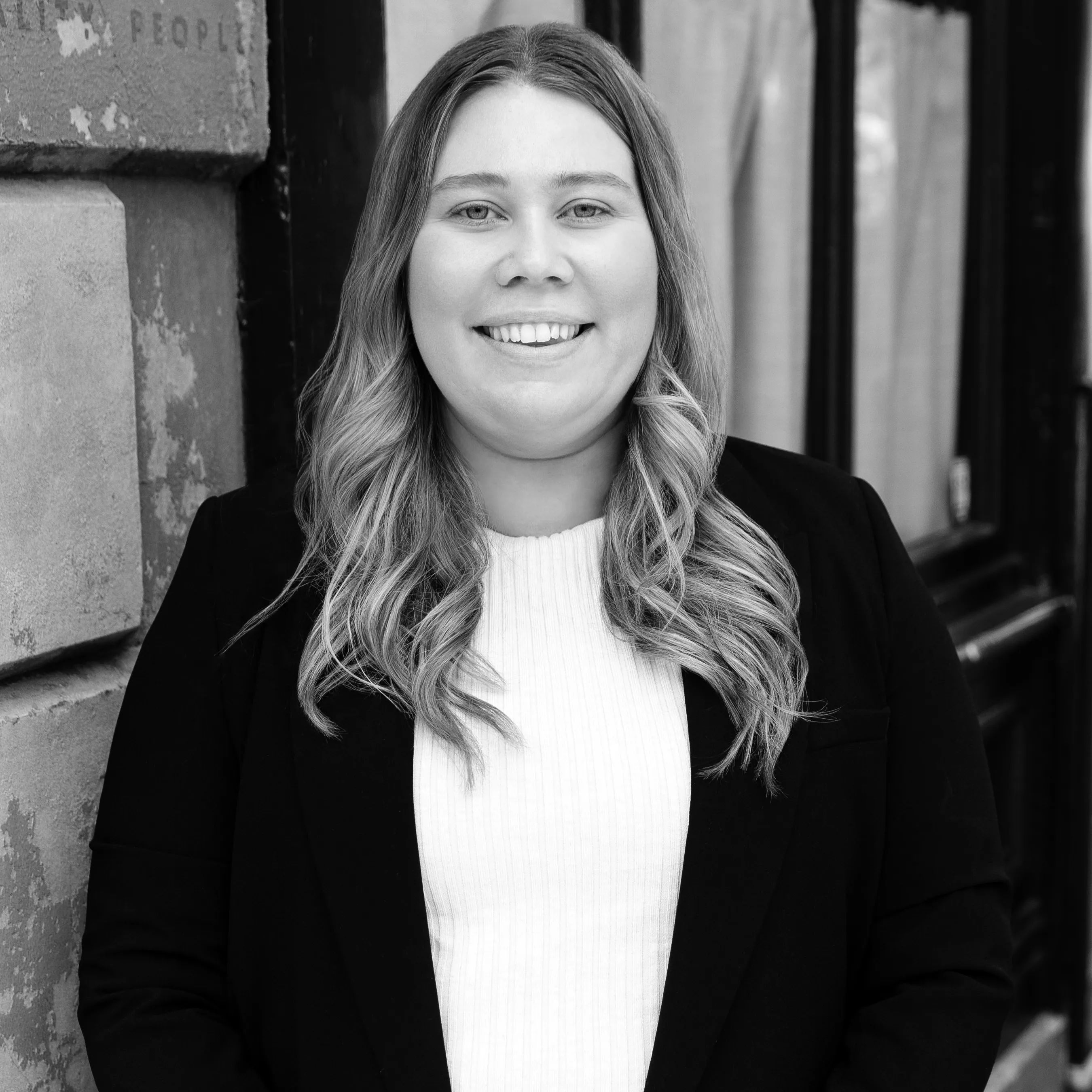 Smiling woman with long wavy hair wearing a black blazer and white top standing against a stone wall and window.