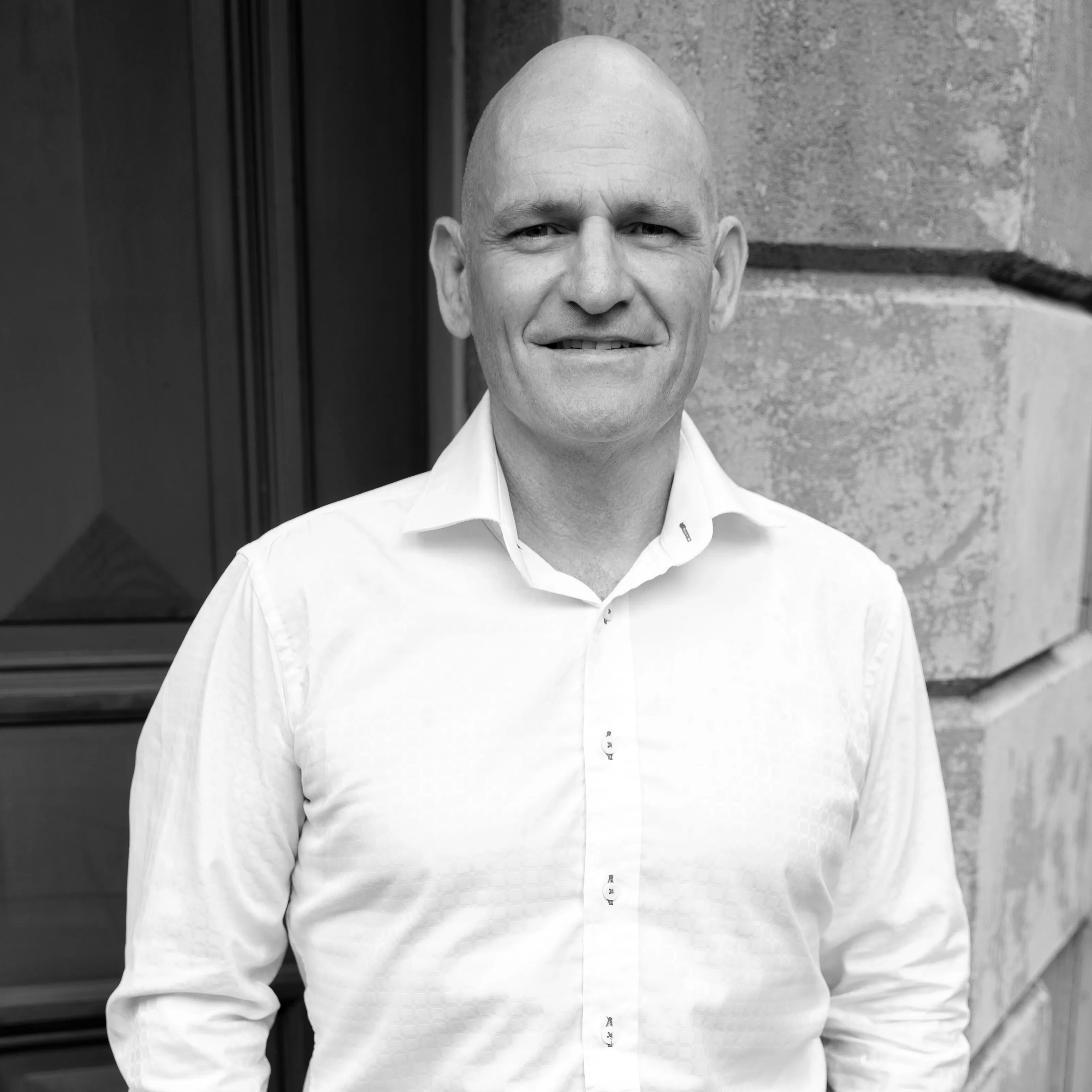 Smiling bald man in a white buttoned shirt standing outside near a textured stone wall.