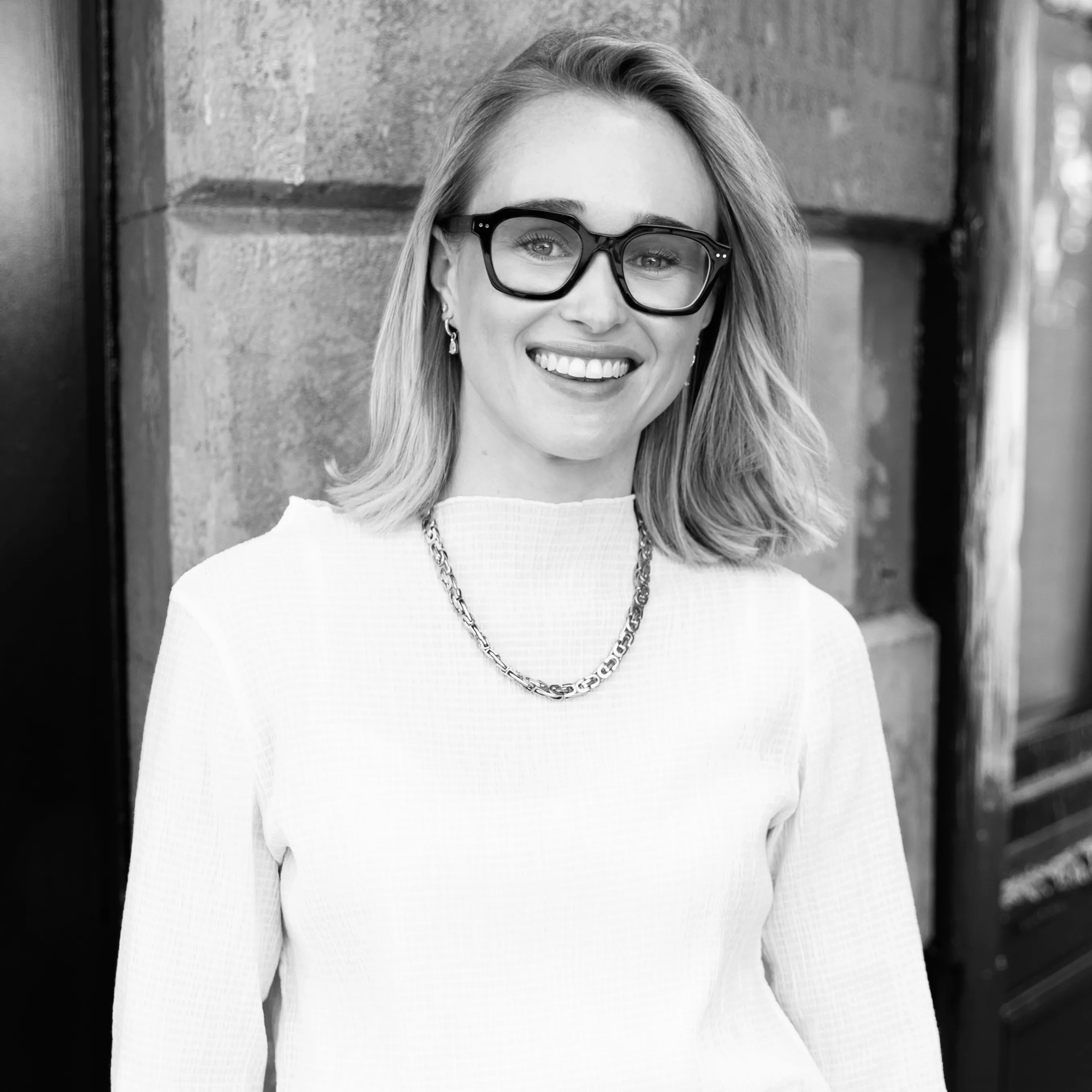 Smiling woman with shoulder-length hair wearing large square glasses, a chain necklace, and a textured white top standing near a stone wall.