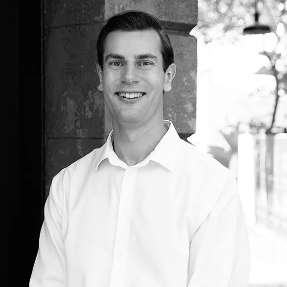 Smiling man wearing a white shirt standing against a stone wall with an outdoor background.